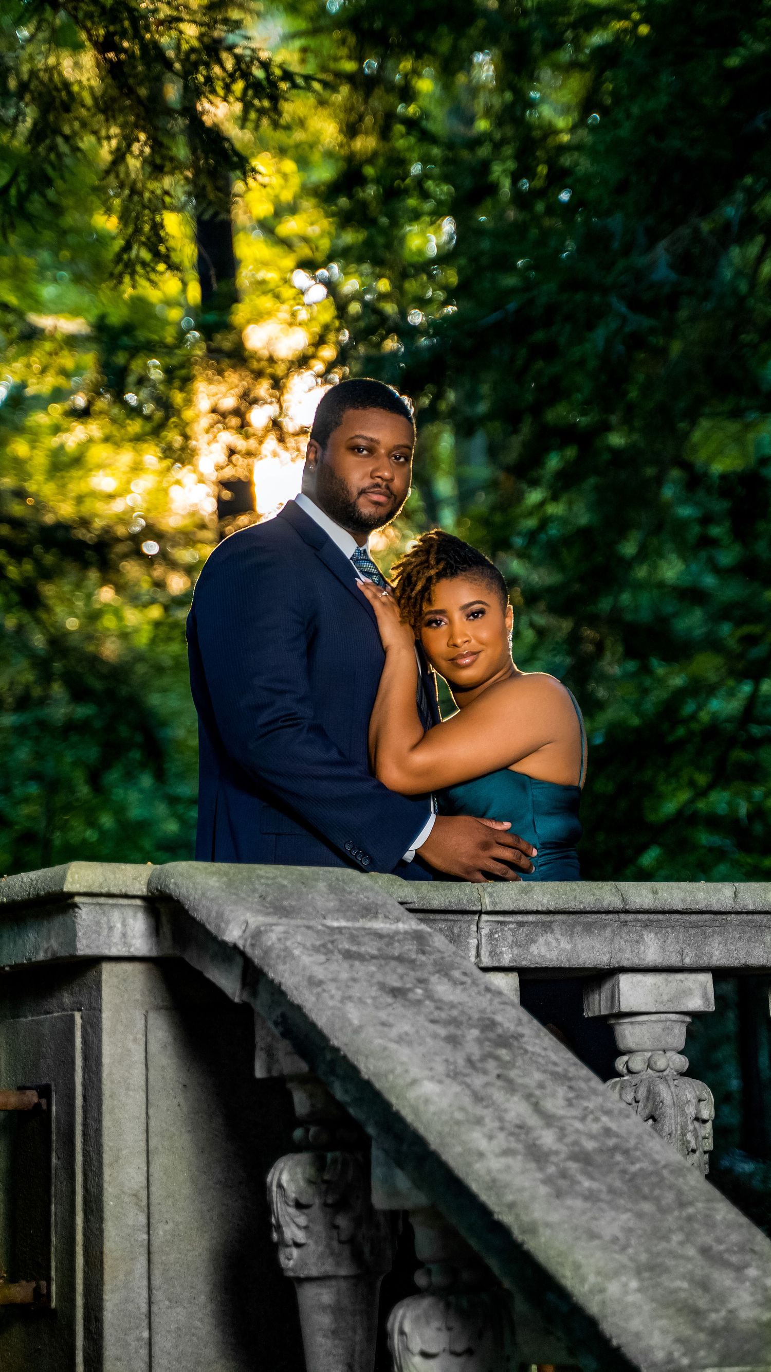 Elegant couple poses on stone balcony surrounded by lush green trees during golden hour engagement shoot.