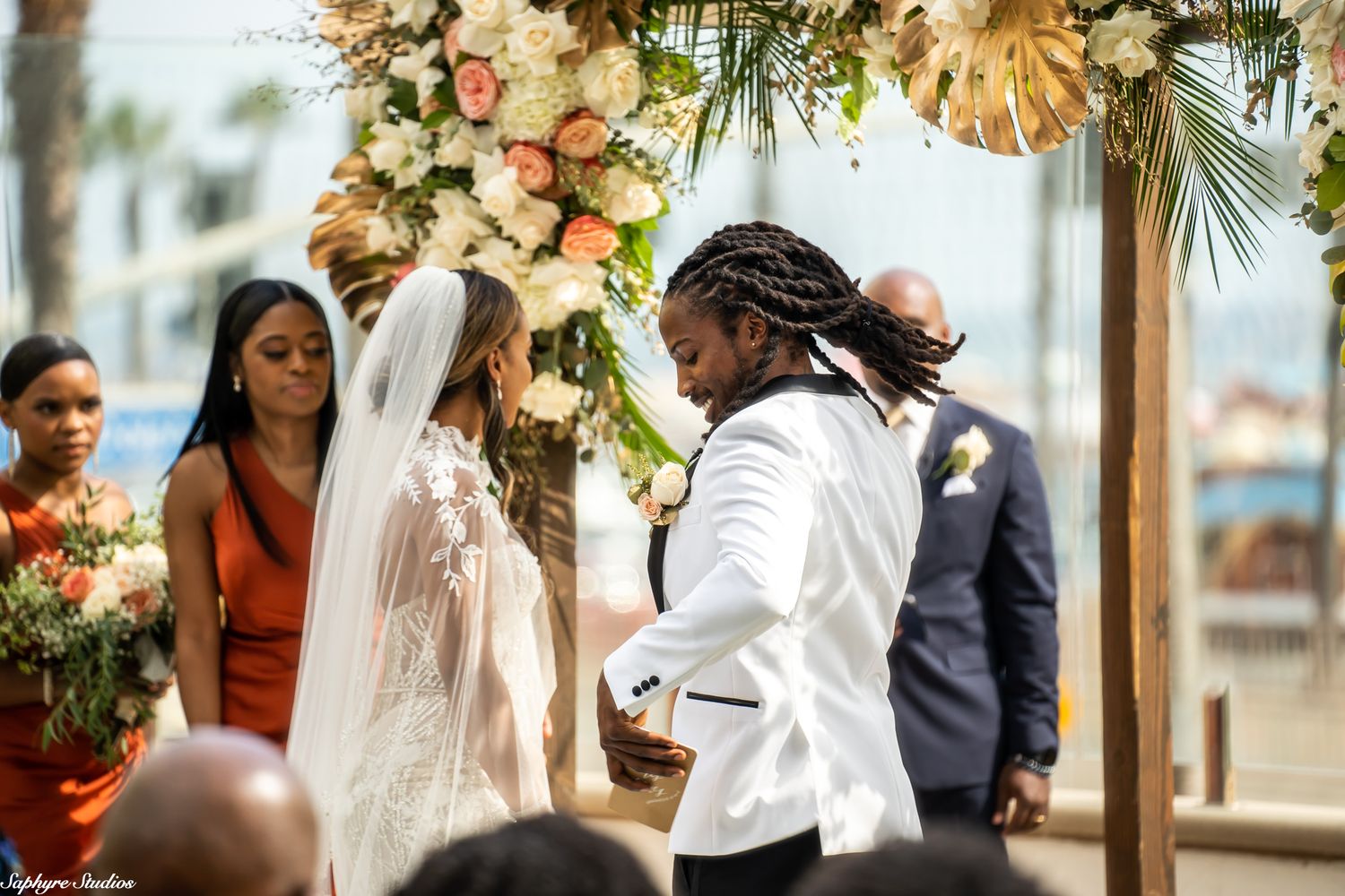 Groom in white suit exchanges rings with bride under floral arch at outdoor wedding.