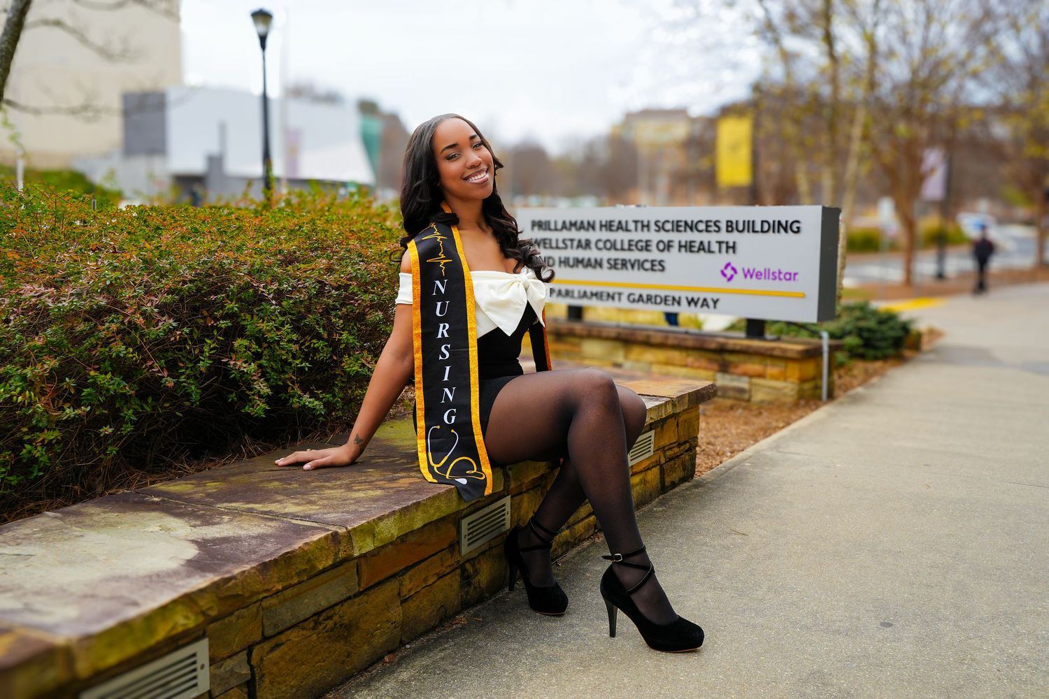 Young African American woman wearing a Nursing graduation sash sitting outside Prillaman Health Sciences Building.