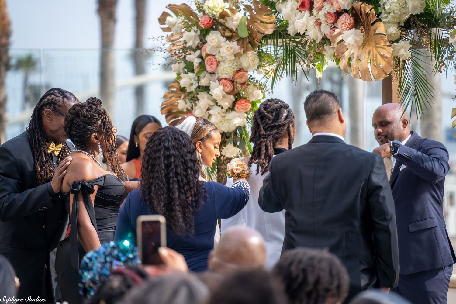Wedding party gathers closely around couple under floral arch during outdoor ceremony.