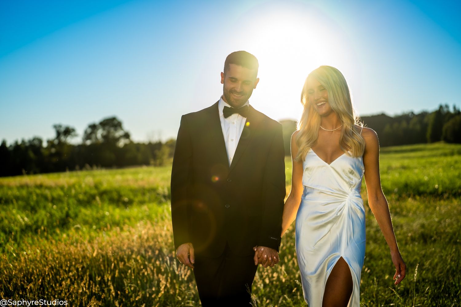 Elegant wedding couple walking hand in hand through a sunlit green field under blue sky.