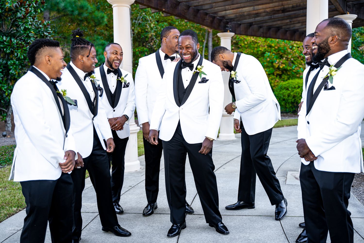 Groom and groomsmen in white and black tuxedos laughing together outdoors at wedding venue.