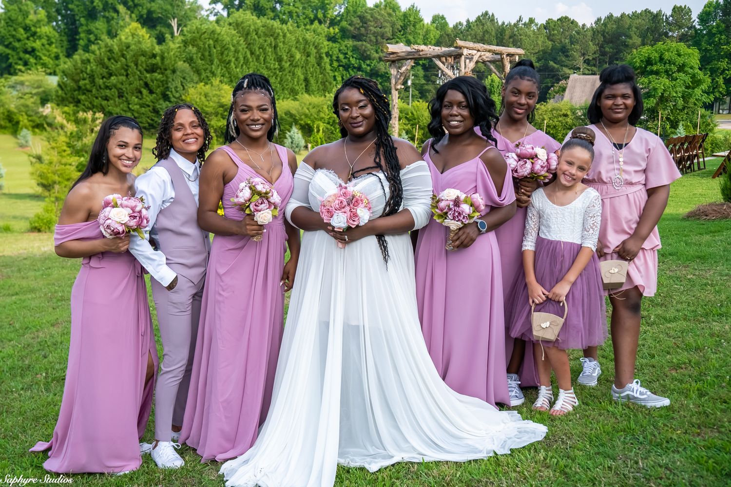 Bride in white gown poses with bridesmaids in dusty rose dresses and flower girl outdoors at garden wedding venue.