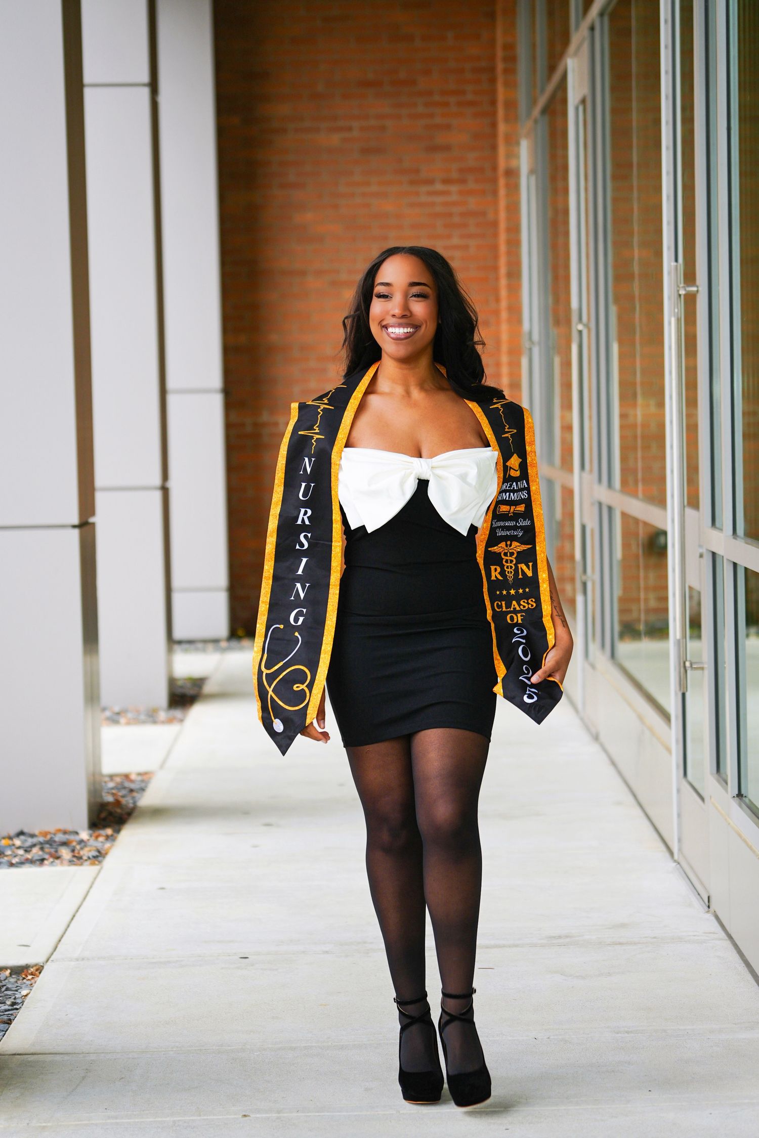 Black woman in nursing graduation stole and black dress walking confidently outdoors.