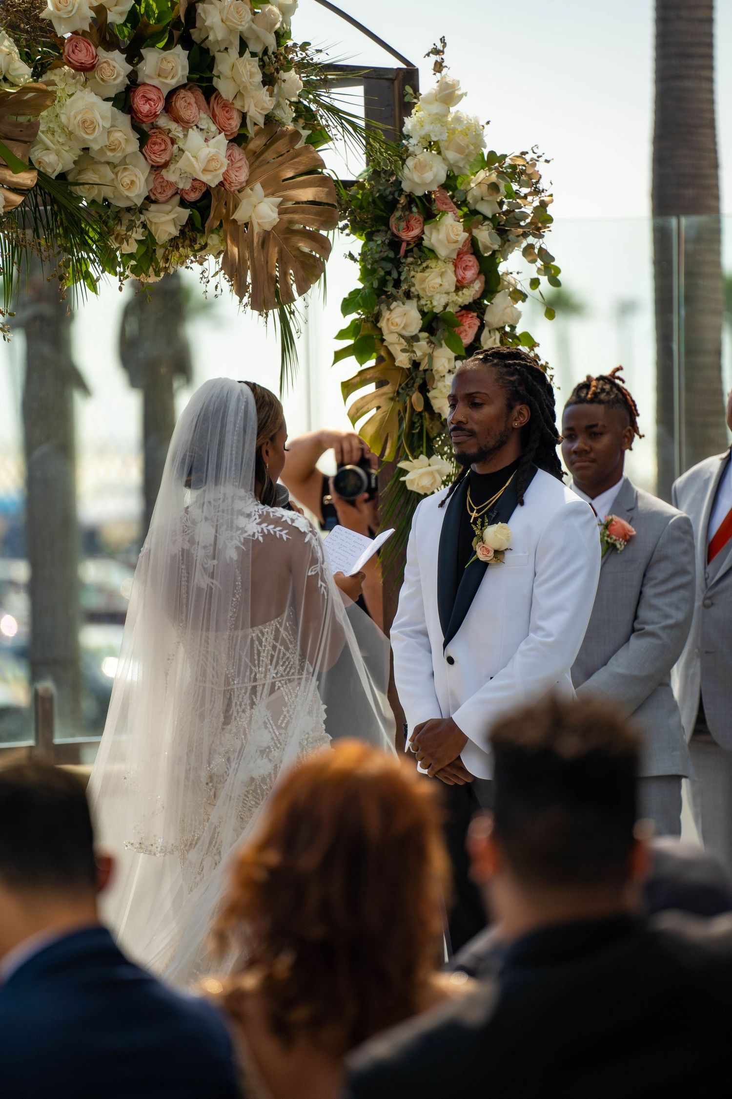 Bride reads vows to groom in white suit under floral arch at outdoor waterfront wedding.