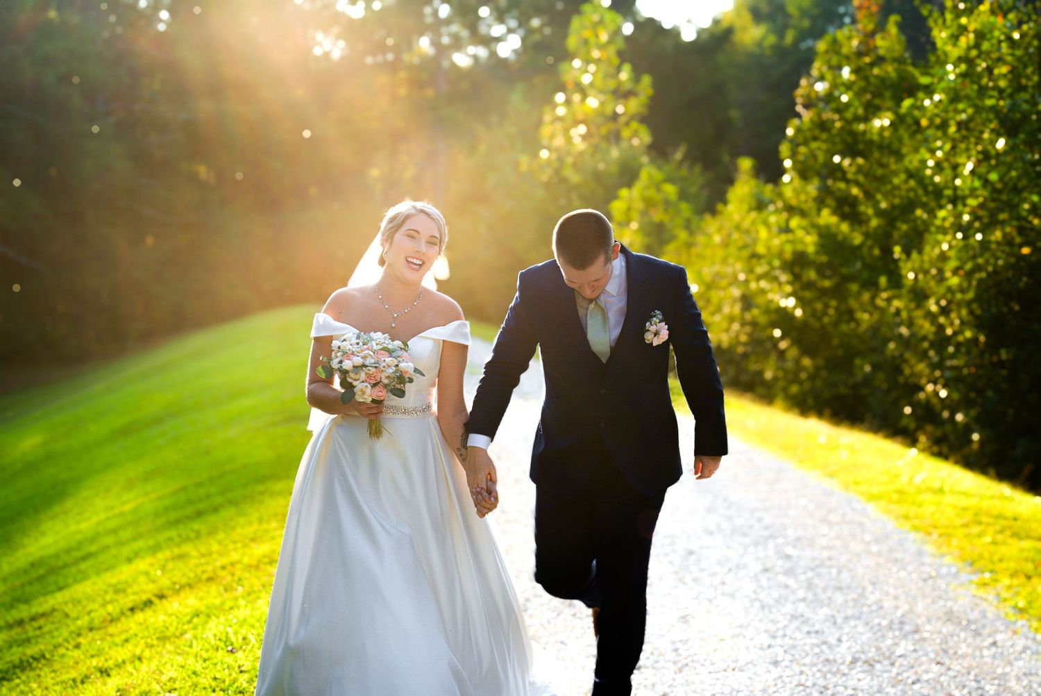 Bride carrying bouquet walks with groom along sunlit garden path during outdoor wedding celebration.