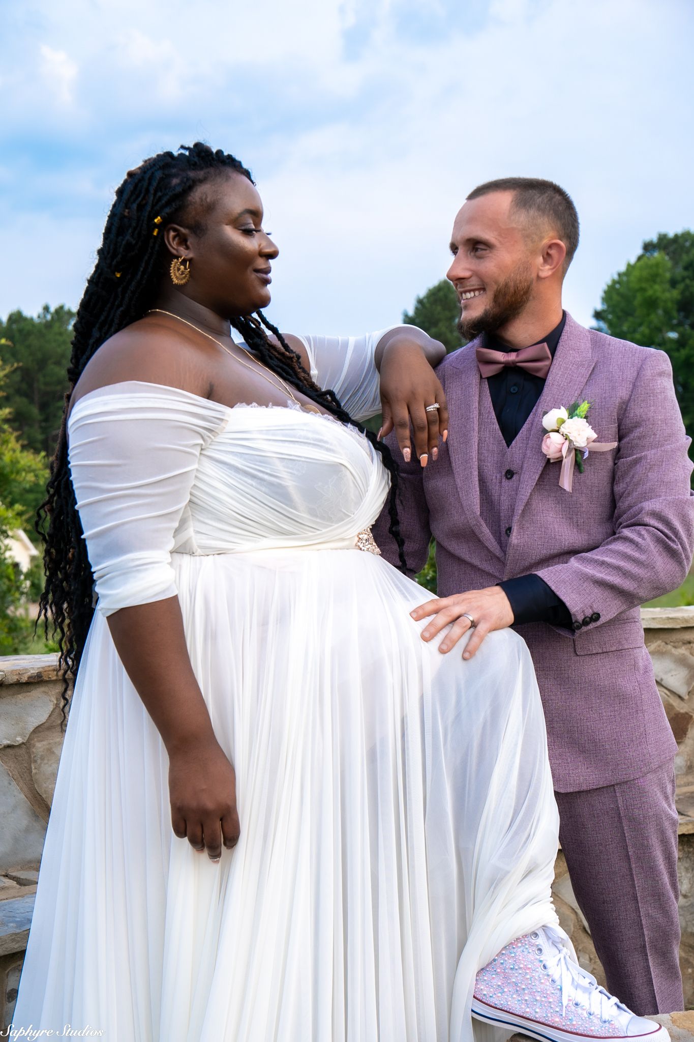 Smiling groom in pink suit stands beside bride in white off-shoulder gown at outdoor wedding.
