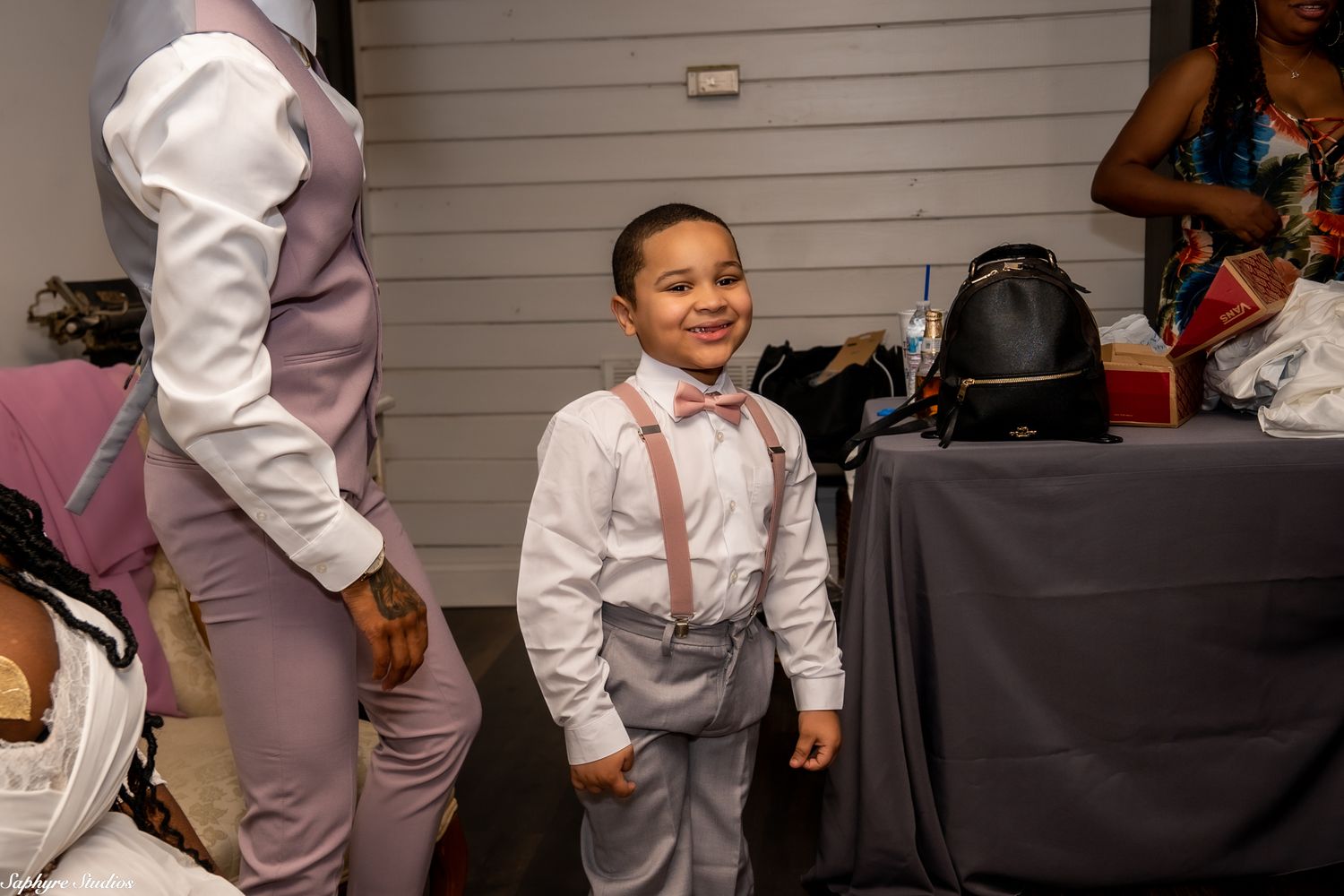 Adorable young boy in white shirt with suspenders and bow tie smiling at wedding reception.