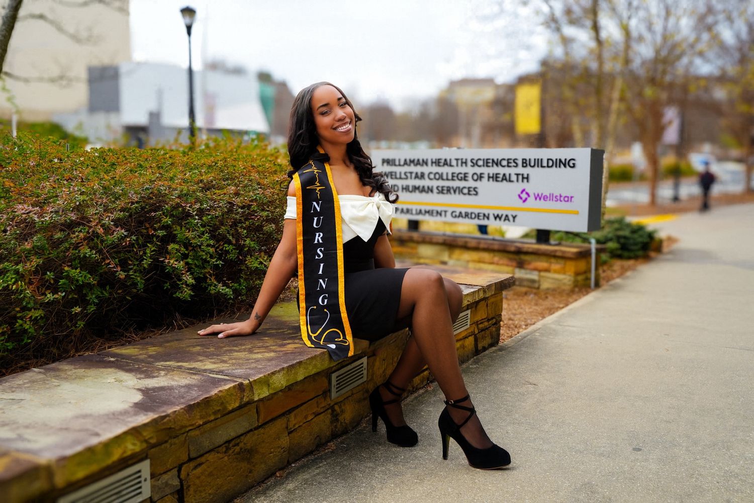Female graduate celebrating in cap and sash outside Wellstar College of Health building.