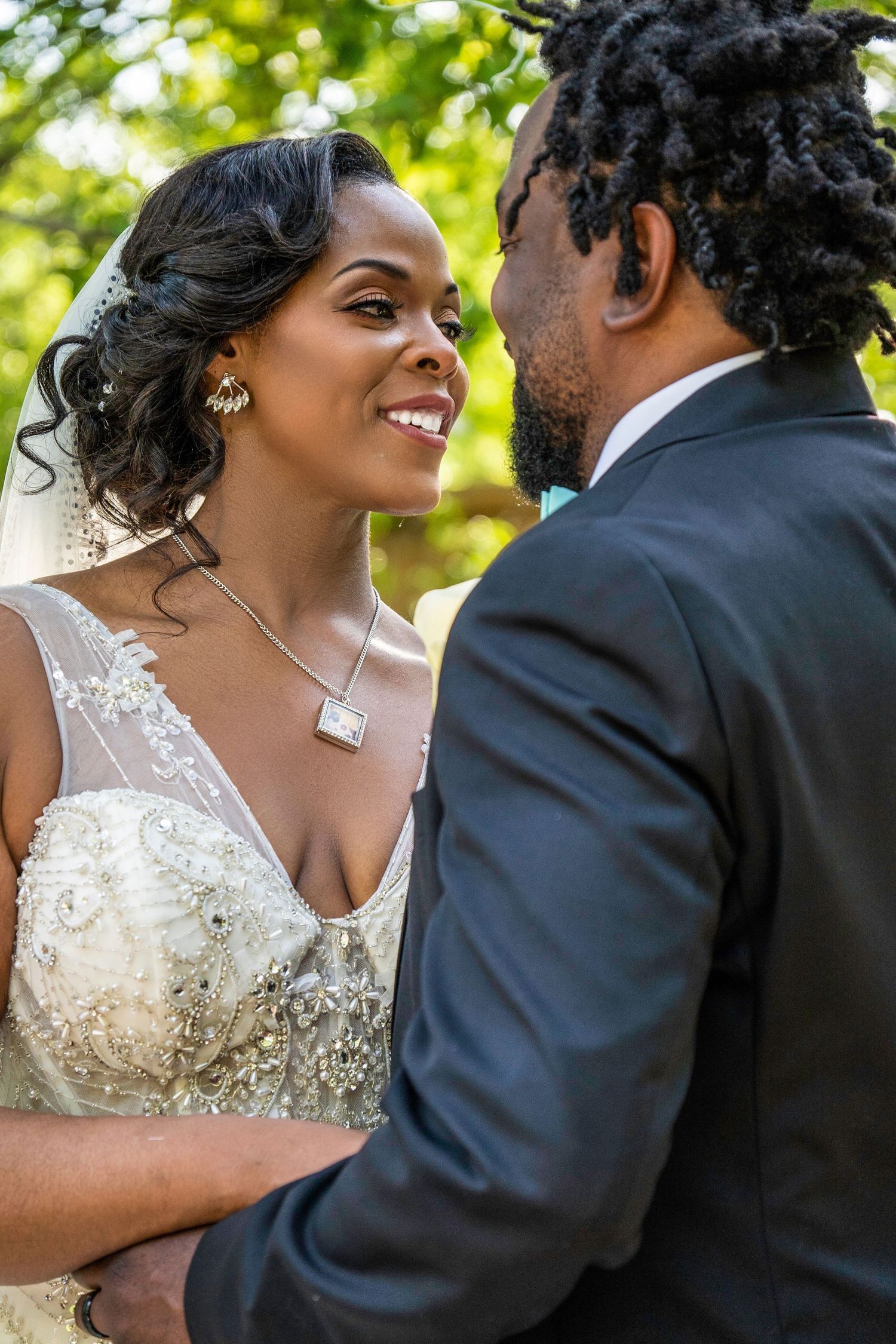 Bride and groom sharing intimate moment outdoors, bride smiling in lace wedding gown with veil, surrounded by green trees.