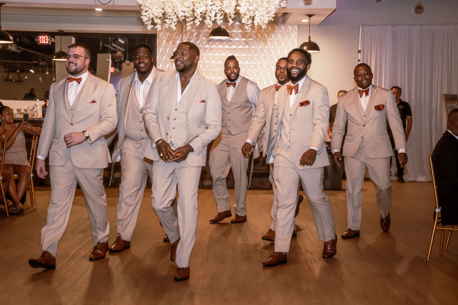 Groomsmen in matching beige suits with bow ties walking confidently across wedding reception dance floor.