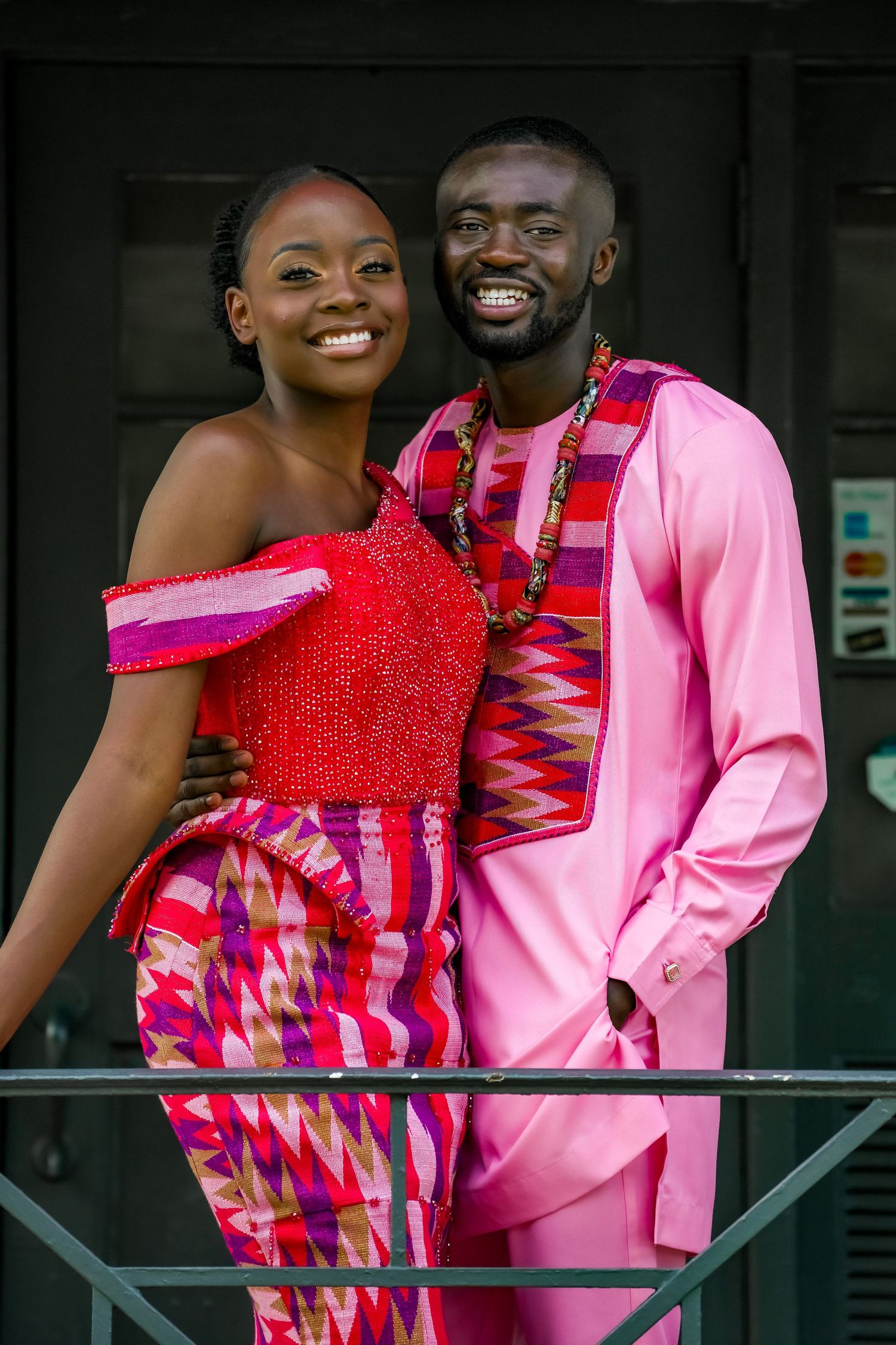 Smiling couple in vibrant traditional African attire pose together at cultural wedding celebration.