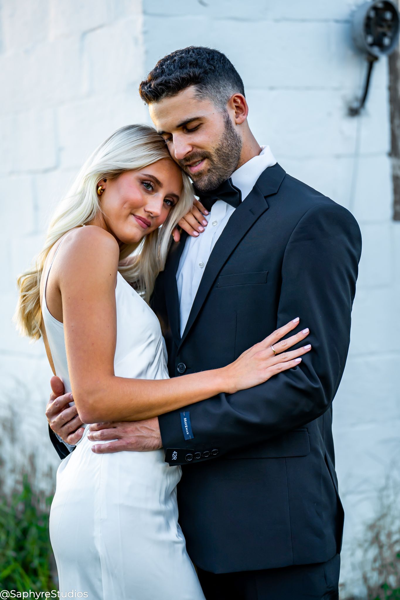 Elegant couple in wedding attire share a tender embrace against a white brick wall.