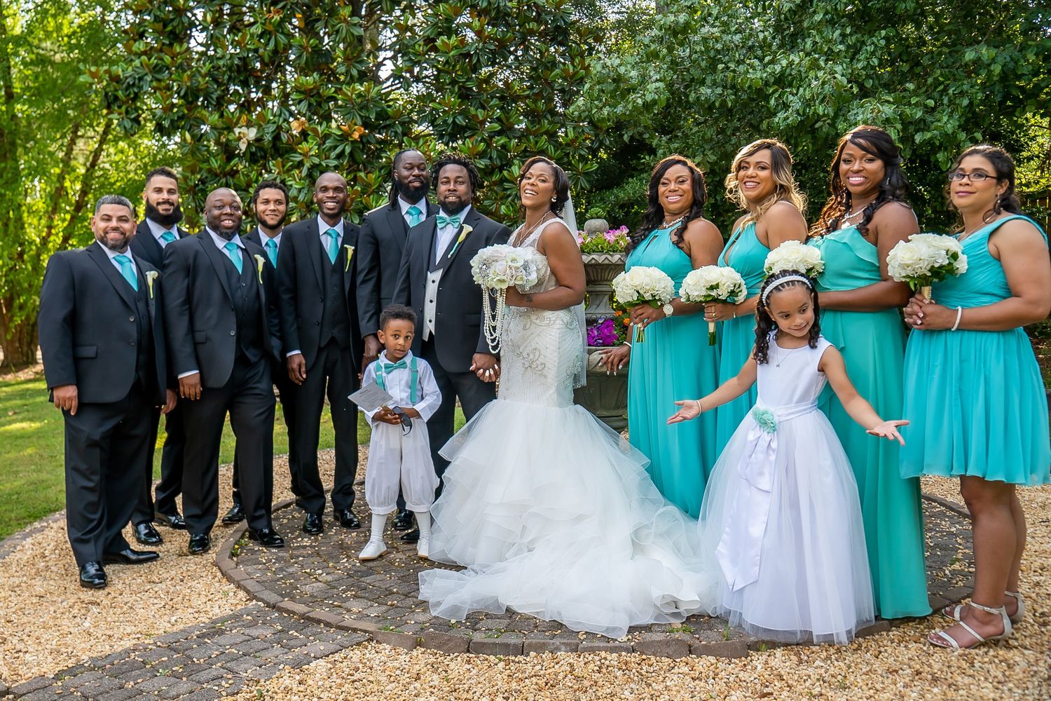 A wedding party poses in teal and black attire with the bride and groom in a lush garden.
