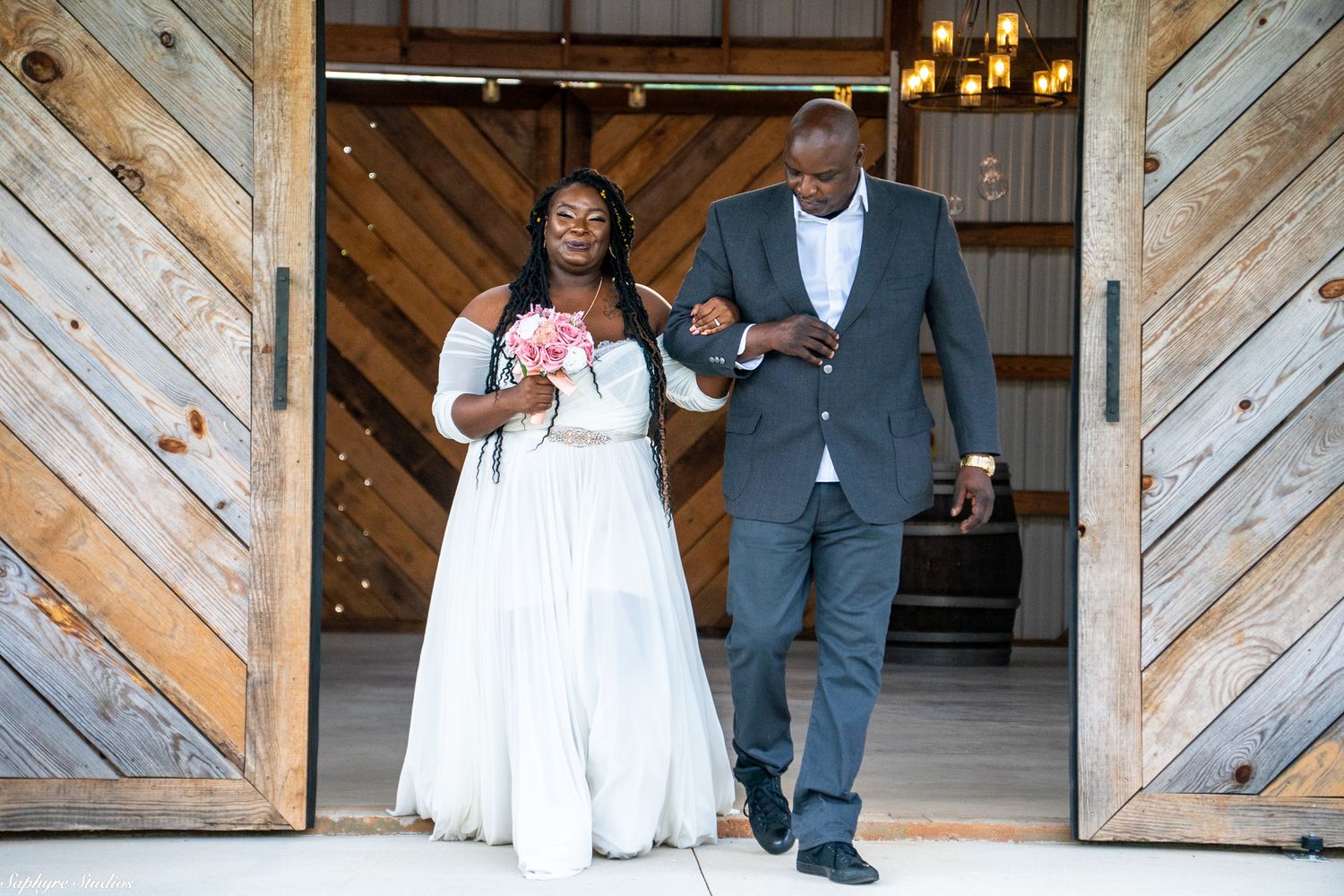 Bride in white gown and groom in gray suit exit barn venue through large wooden doors after ceremony.