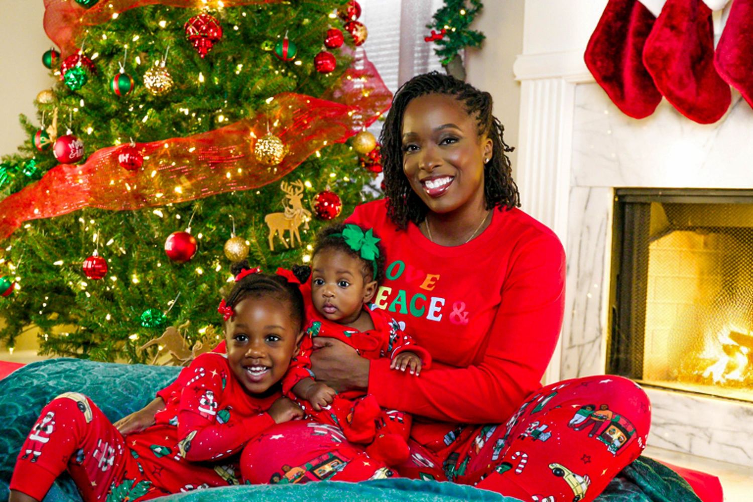 Mother and two young children in matching red Christmas pajamas by decorated tree.