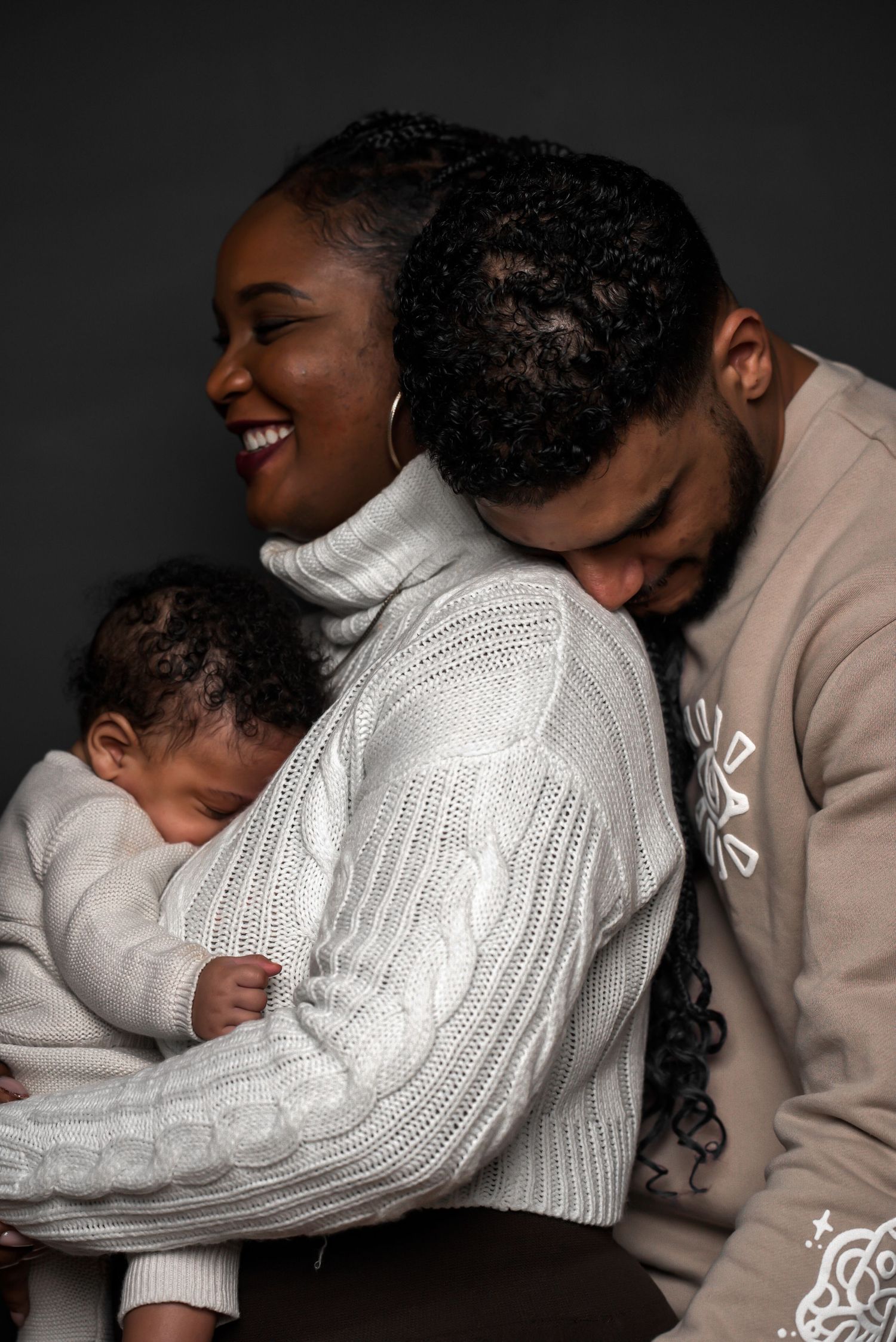 Loving couple holding sleeping newborn baby together, smiling warmly against dark background.