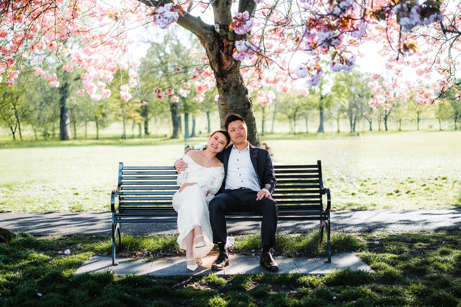 Newlywed couple sitting closely on a park bench under blooming pink cherry blossom trees in spring.