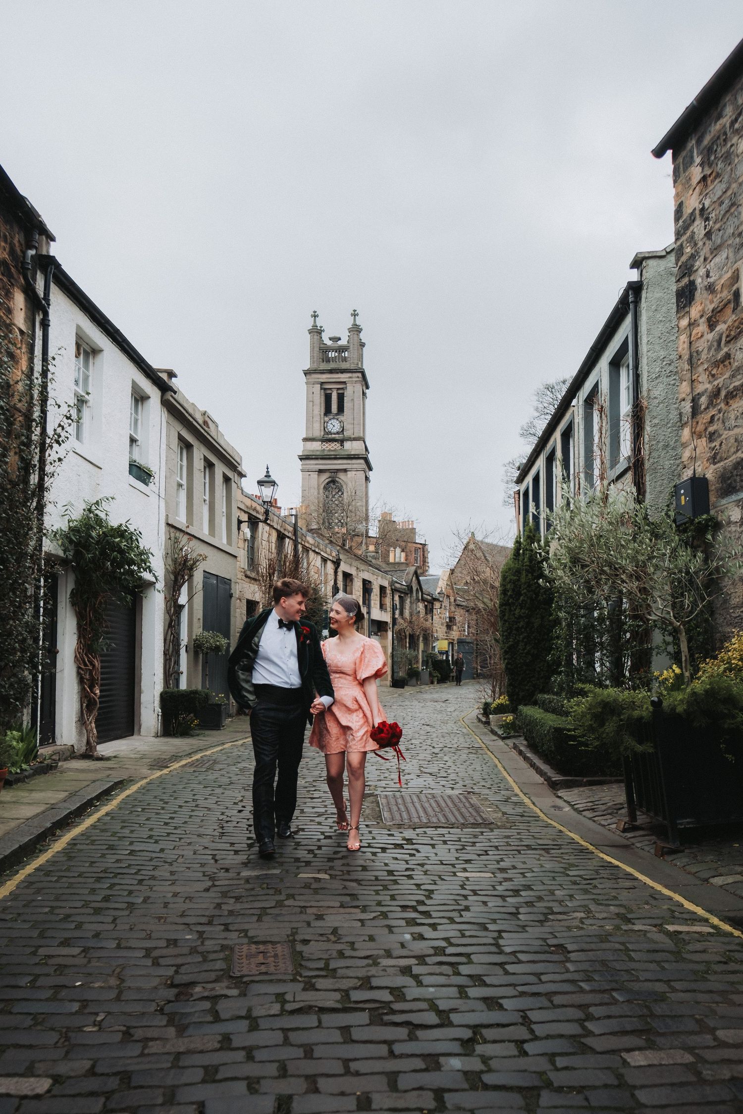 Couple walking arm in arm on cobblestone street with historic clock tower backdrop on cloudy day.