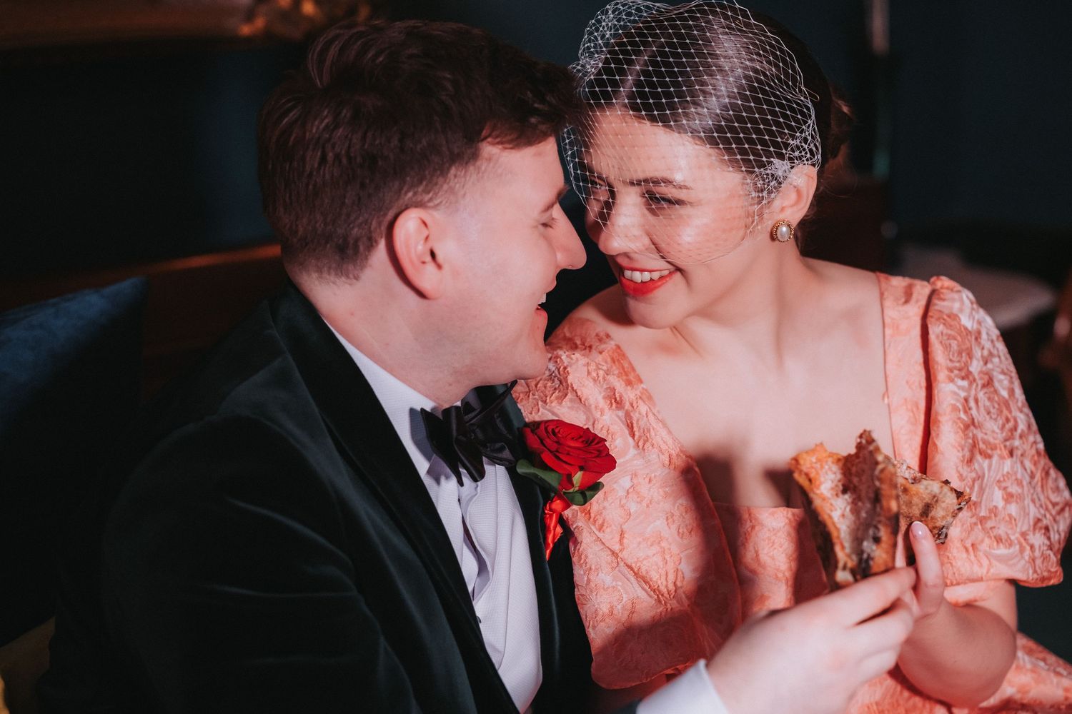 Bride in pink lace dress and groom in tuxedo sharing a romantic moment with red rose indoors.