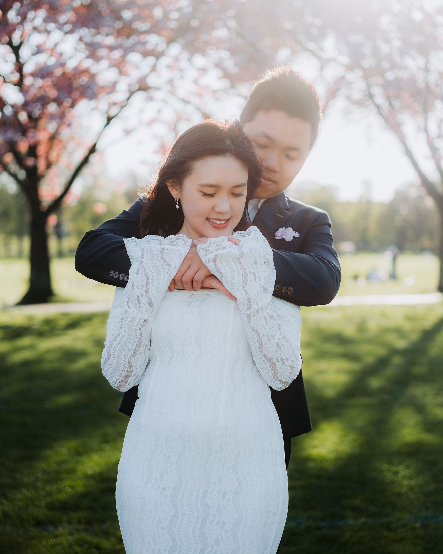Groom embracing bride from behind under cherry blossom trees in warm golden evening light.