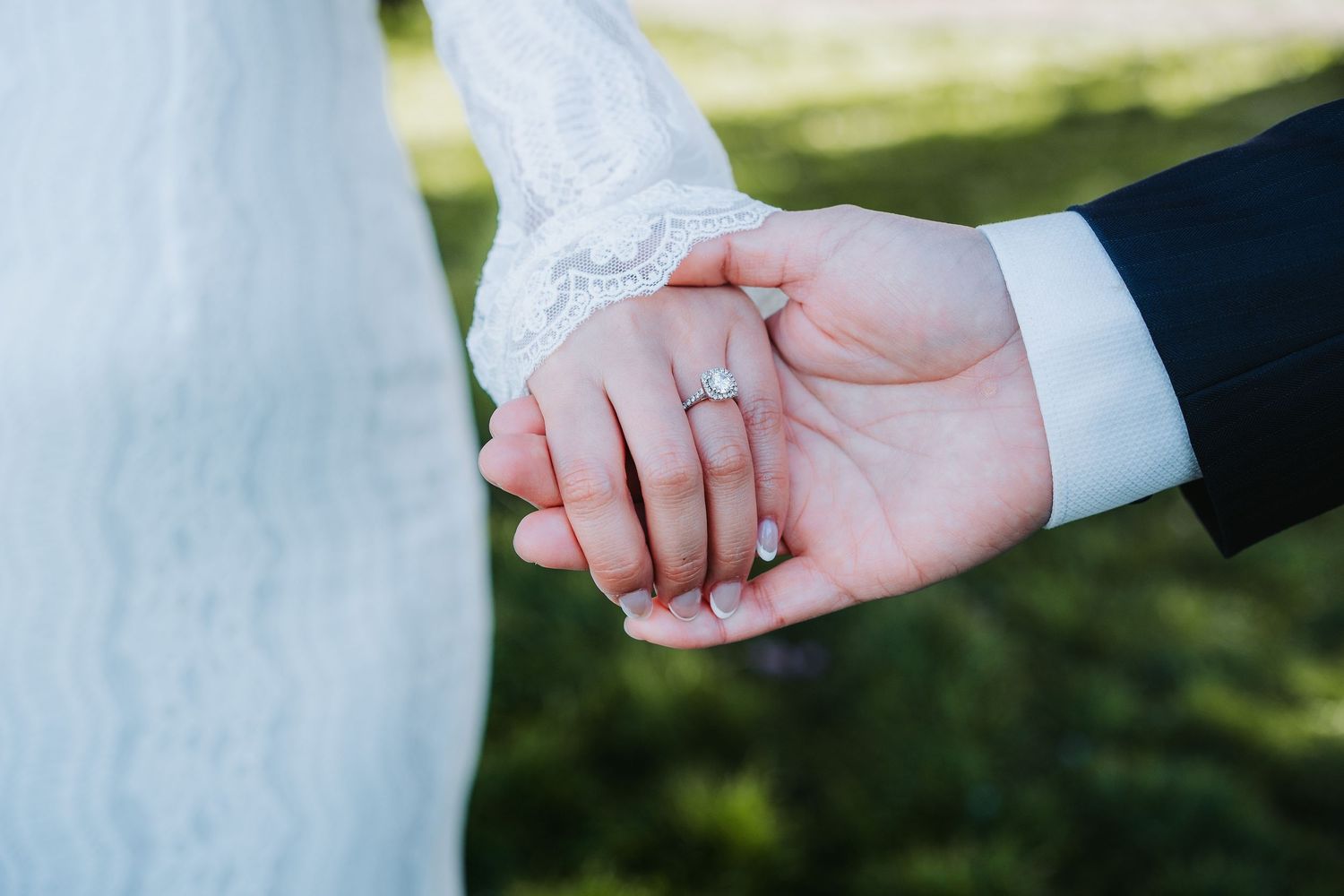 Close-up of bride and groom holding hands showing diamond engagement ring with lace wedding dress.