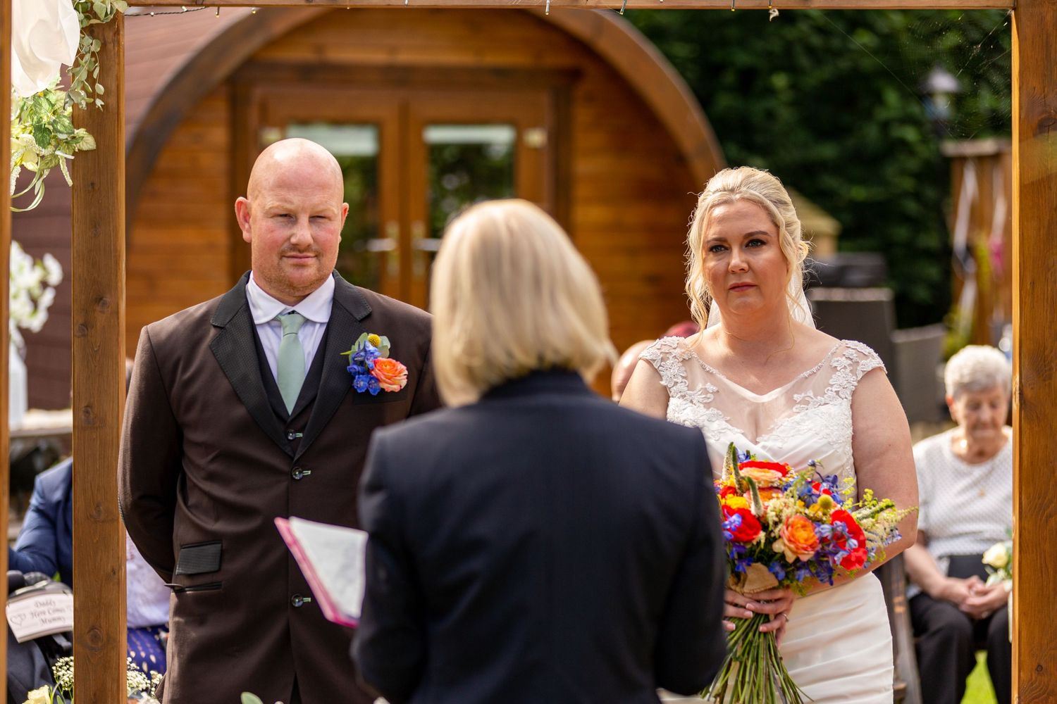Outdoor ceremony at The Gables Escomb; couple stand before celebrant with colorful bouquet and rustic backdrop.