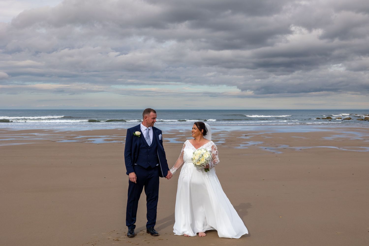 Couple hold hands on Tynemouth beach; moody sky and waves create a romantic coastal scene.