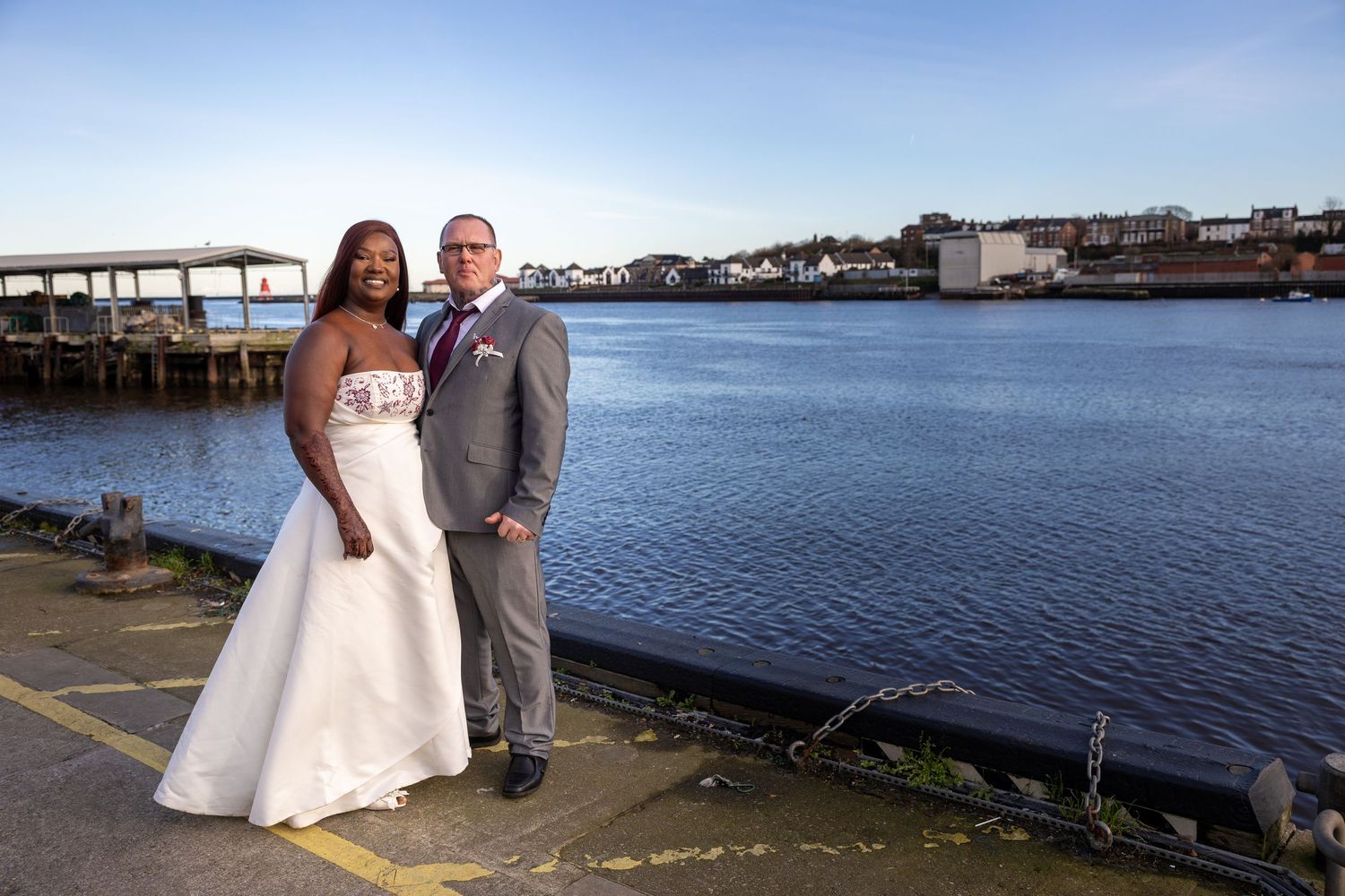Couple pose by waterfront in Tynemouth; calm sea and harbour create a scenic backdrop.