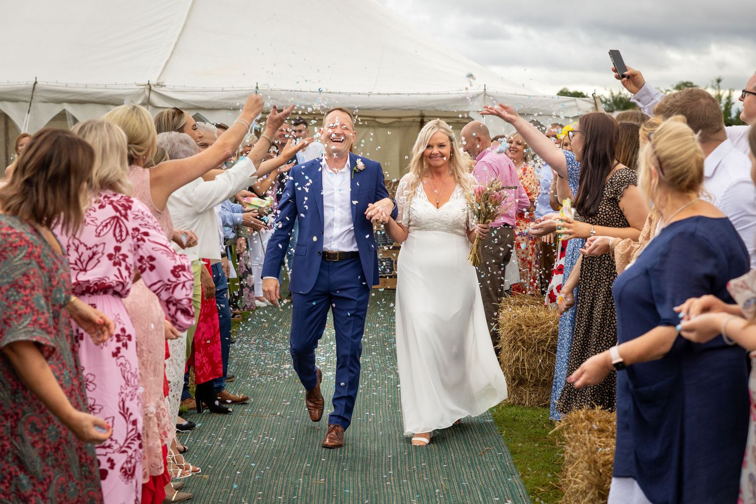Newlyweds walk through confetti tunnel; guests cheer along outdoor aisle, joyful celebration moment.