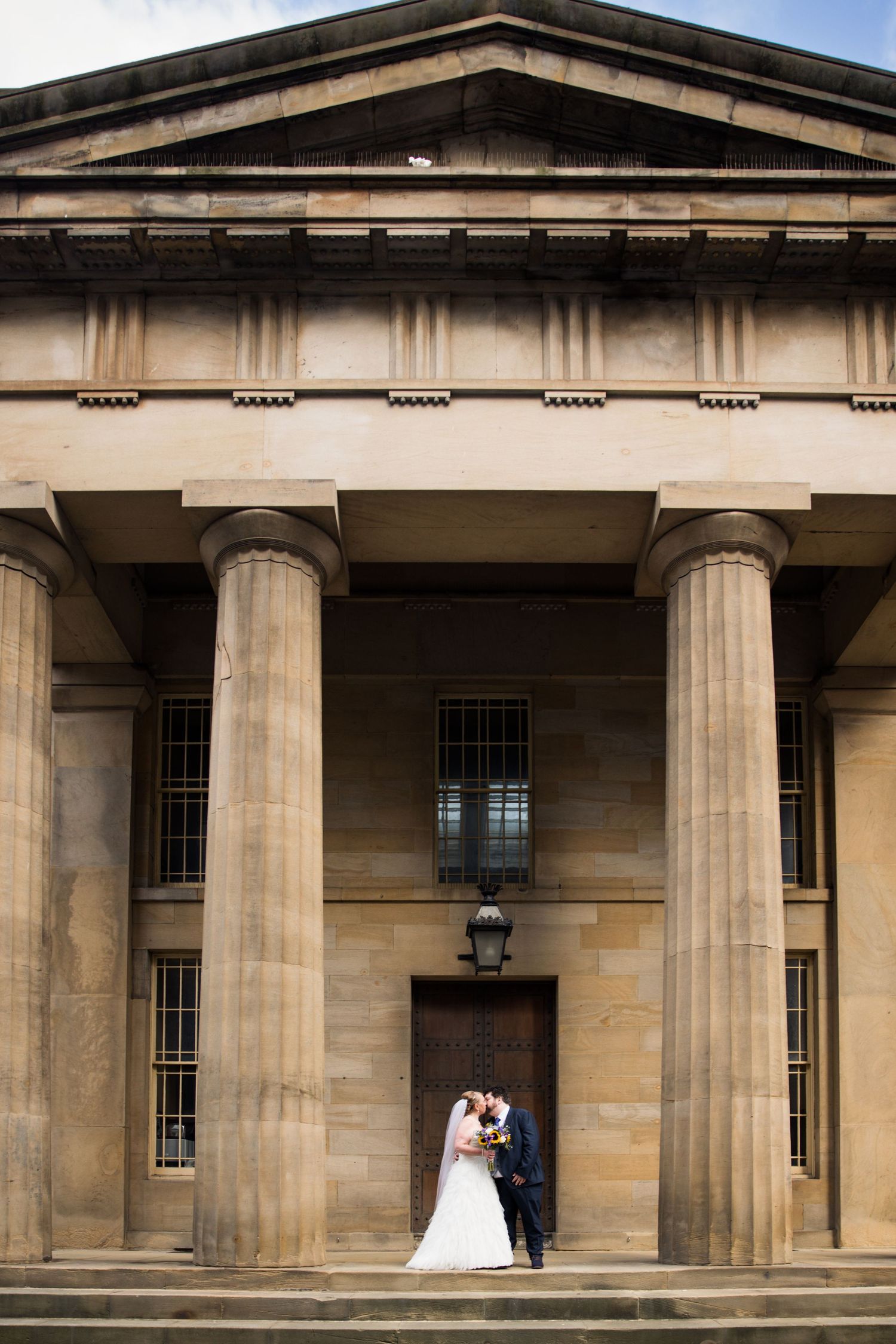 Couple stand beneath grand columns at The Vermont Hotel; striking architecture frames elegant portrait.