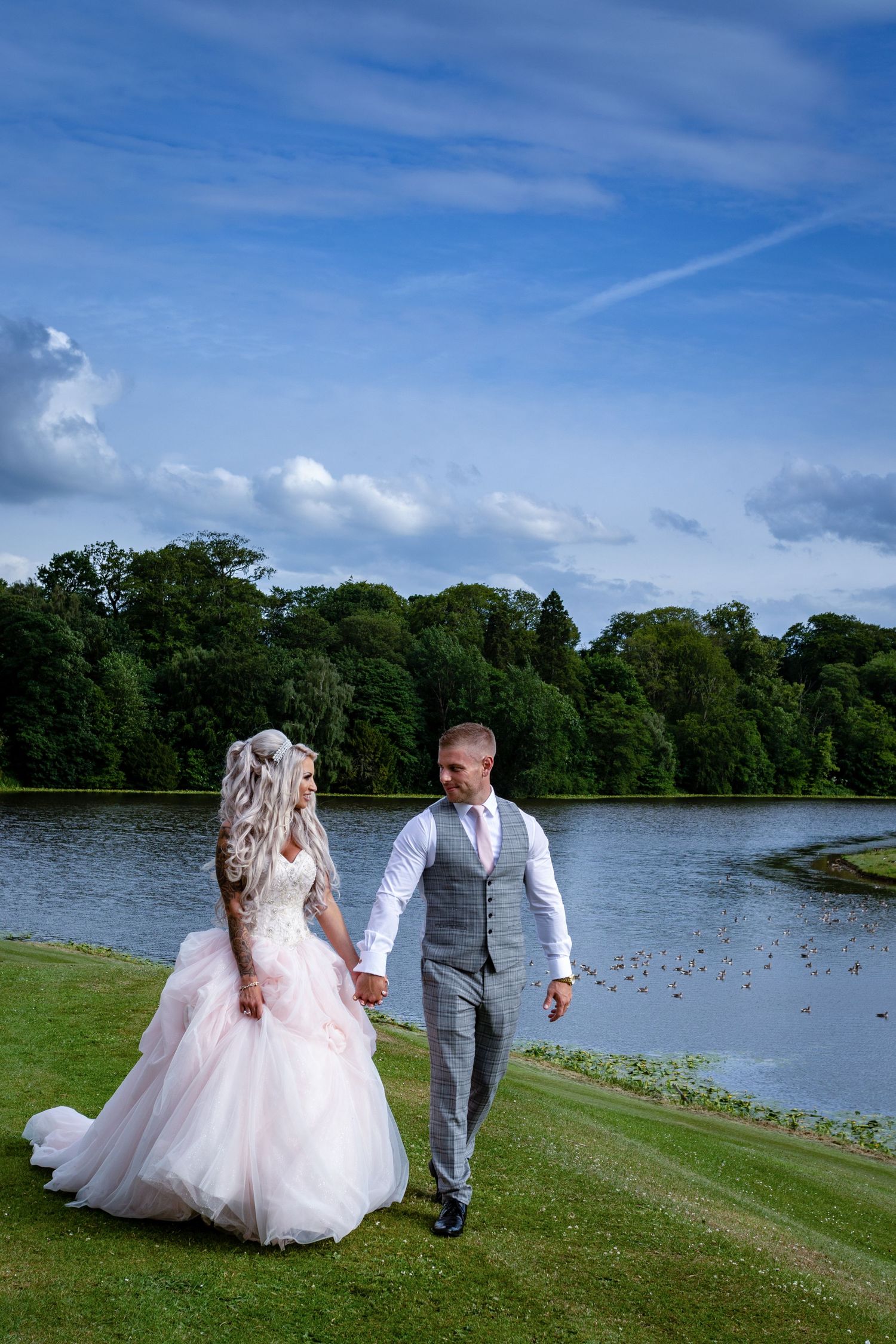 Couple walk by lake at Wynyard Hall; peaceful setting with water, greenery, and soft evening light.