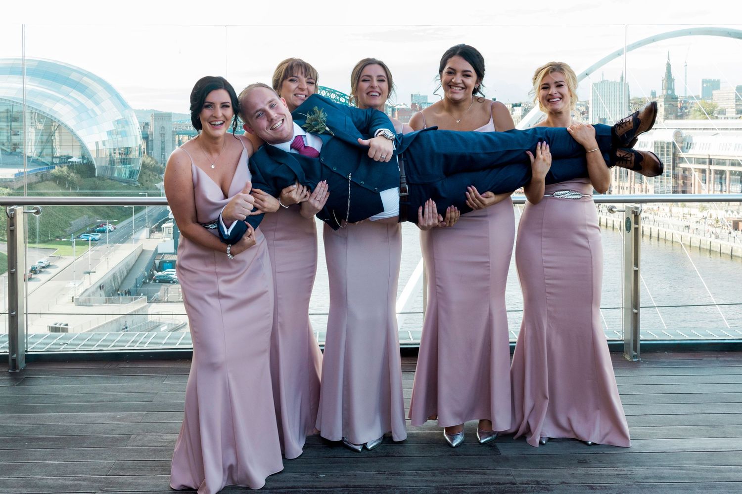 Bridesmaids lift groom at Baltic Newcastle, fun wedding photo with Tyne Bridge views, capturing stylish North East UK wedding moments