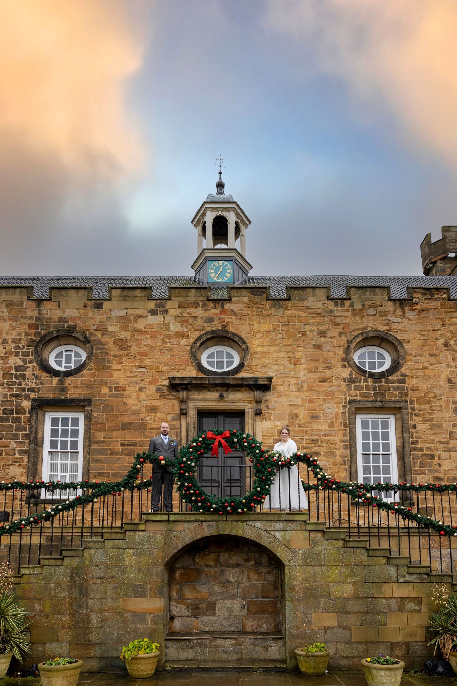 Couple stand on balcony at Lumley Castle; festive decor and historic stone backdrop.