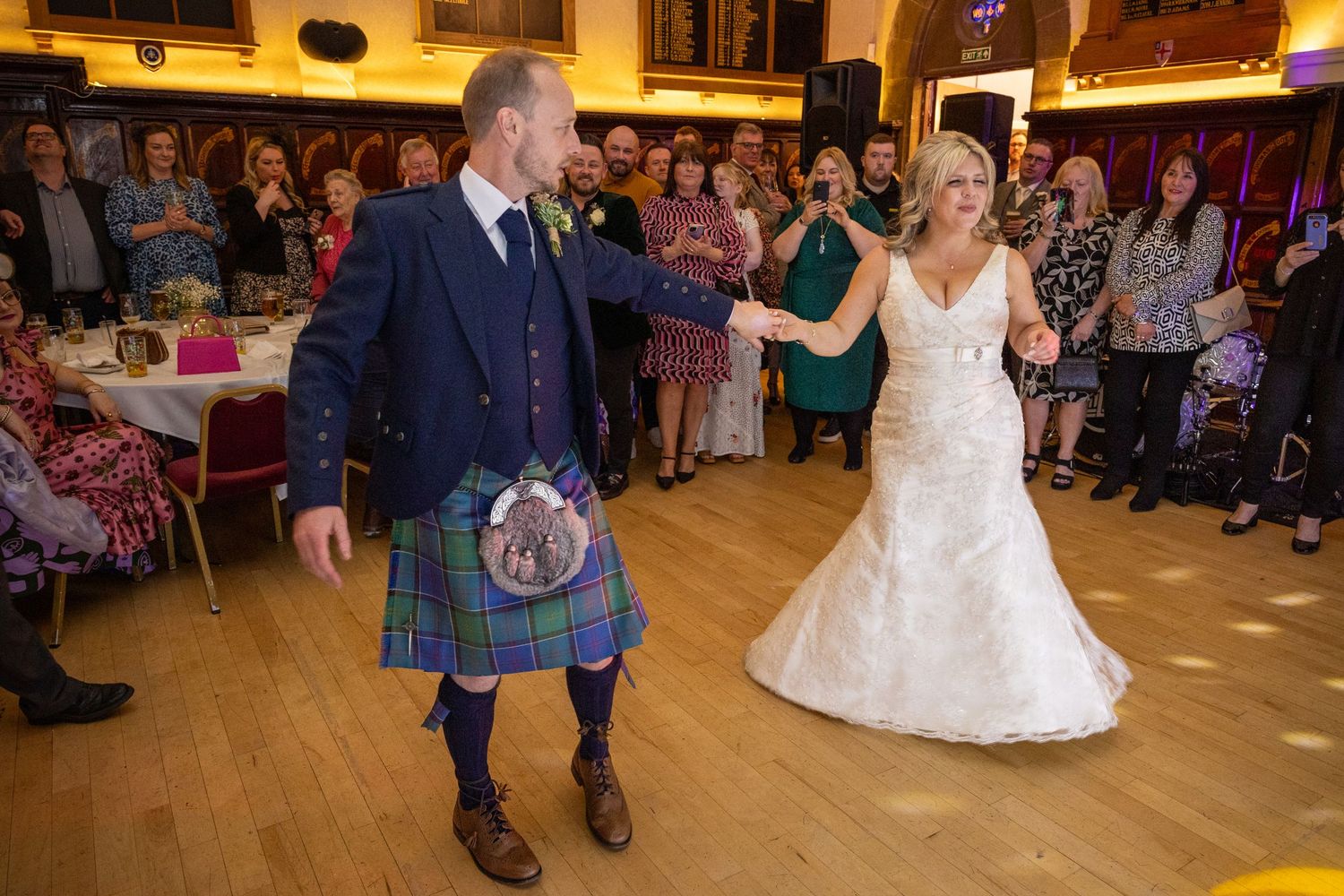 Bride and groom ceilidh dance, groom in kilt, lively wedding reception, guests watching, fun Scottish wedding celebration