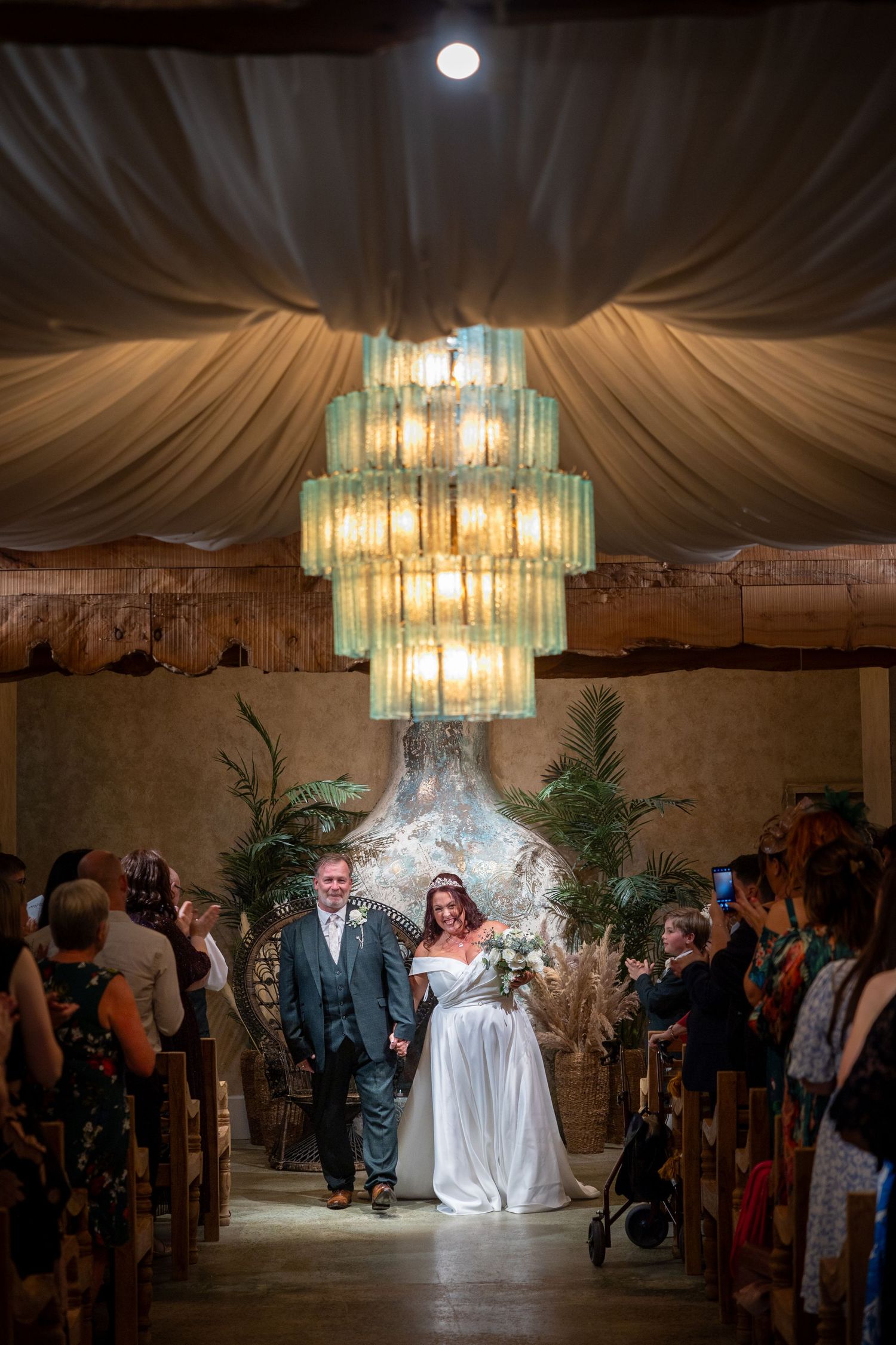 Bride and groom walk down aisle at Runa Farm, elegant indoor ceremony with chandelier, joyful wedding moment captured
