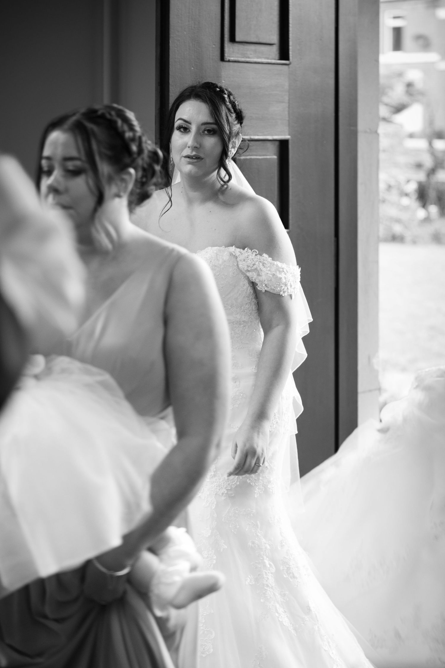 A candid, black-and-white moment as the bride pauses by the doorway, watching her bridal party prepare—quiet anticipation before the ceremon