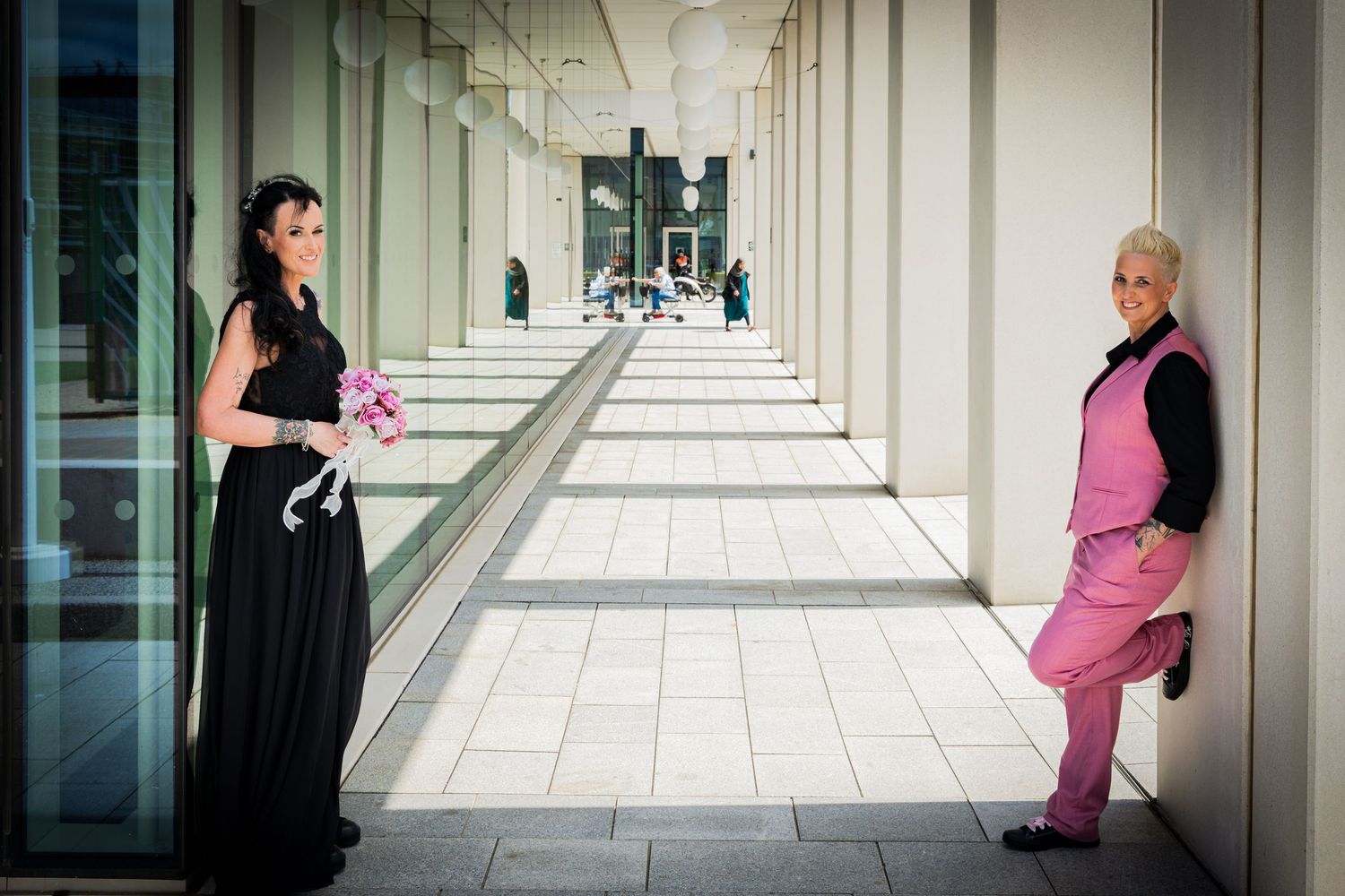 Wedding couple posing beneath the colonnade at Sunderland City Hall, one wearing a black dress and the other a pink suit.