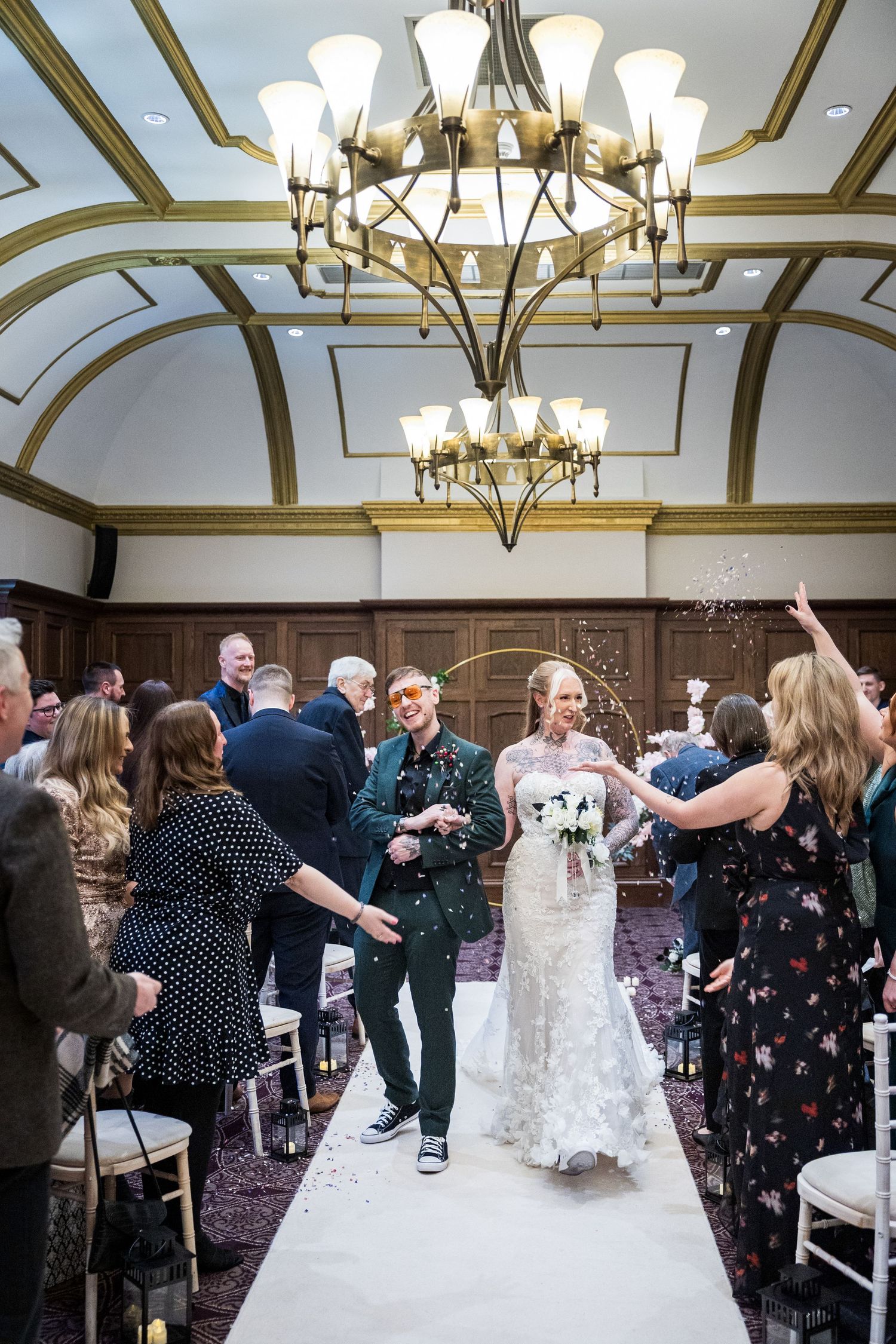 Confetti toss at The County, Newcastle; couple walk aisle as guests celebrate beneath grand chandelier.