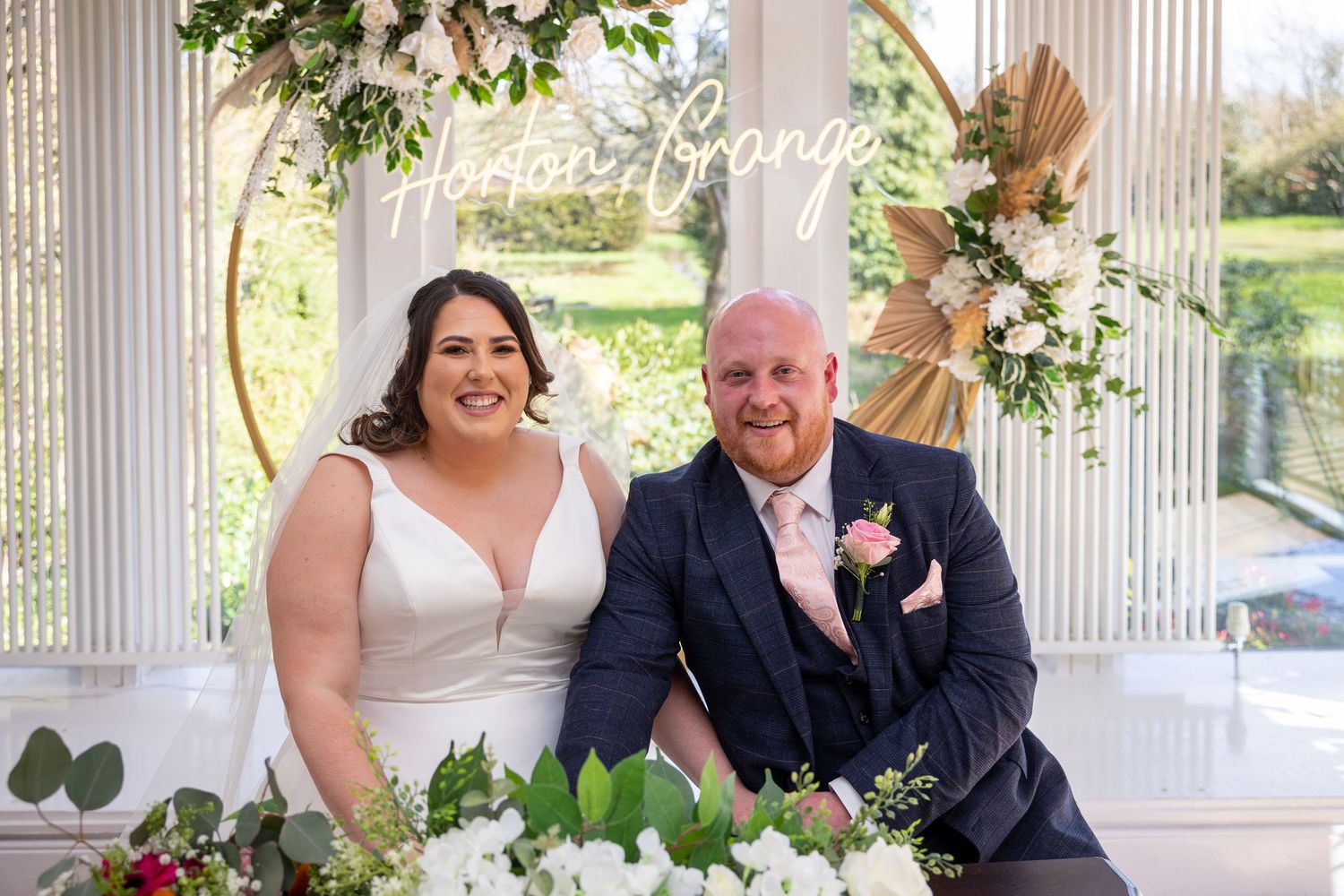 Couple seated at floral ceremony backdrop; bright, modern setting with greenery and soft, romantic styling.