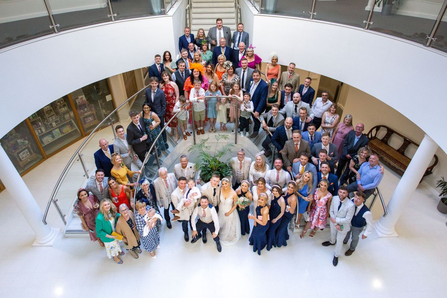 Large wedding group photo indoors, guests gathered on staircase, fun overhead perspective, modern venue, lively and colourful celebration mo