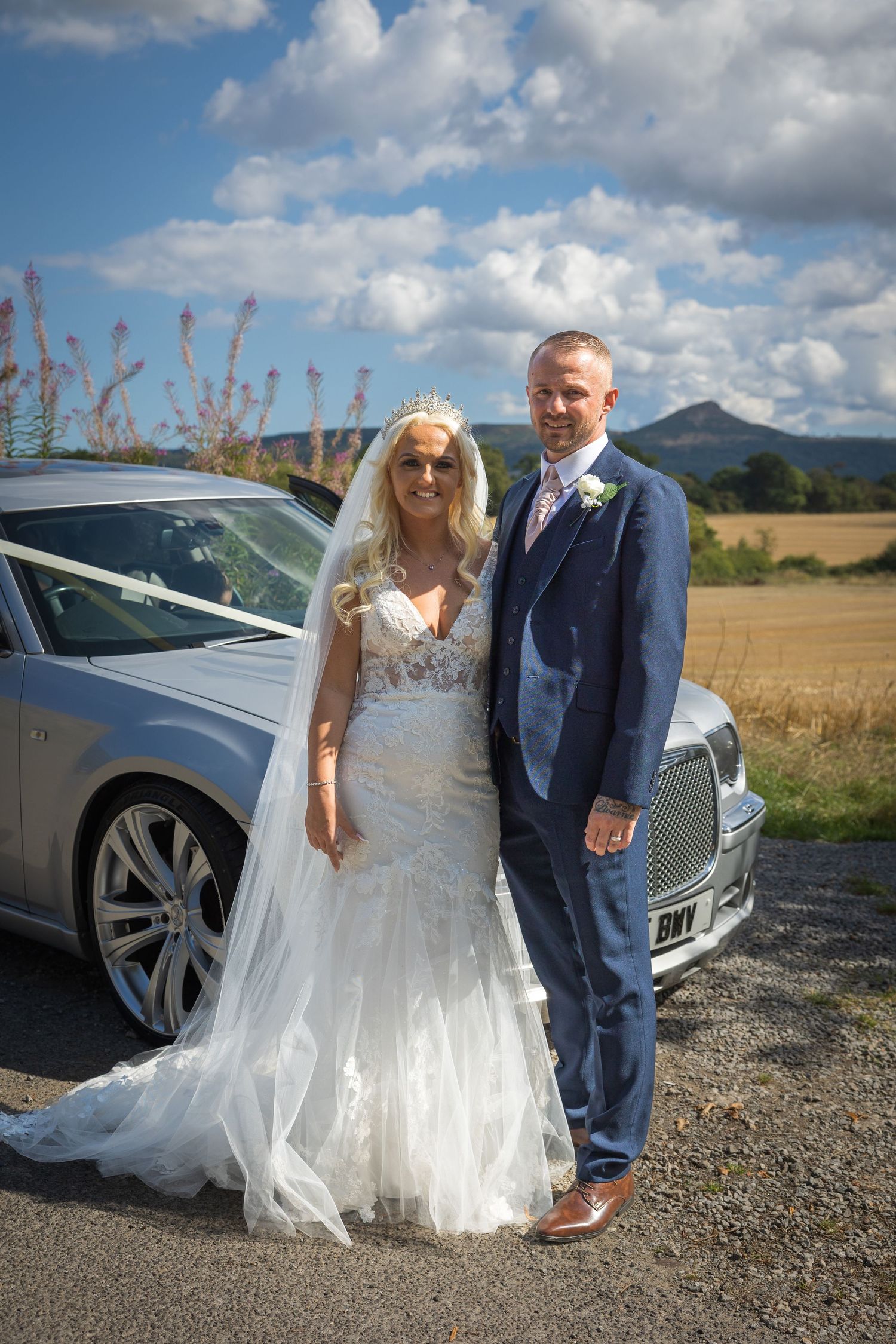 Bride and groom by wedding car with Roseberry Topping in background, scenic countryside wedding, blue skies, romantic UK outdoor portrait