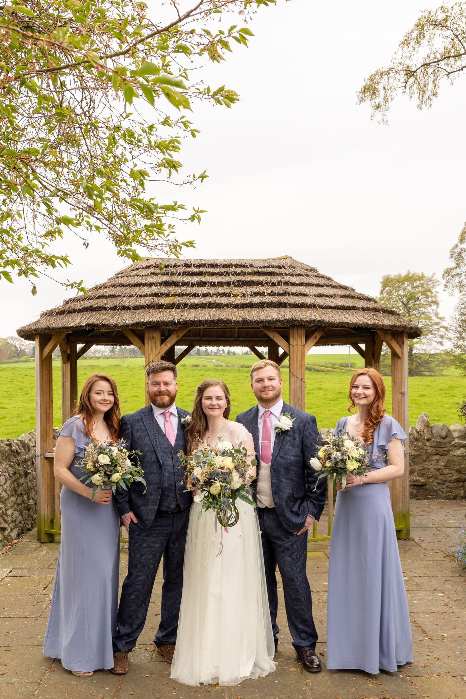 Wedding party pose under wooden gazebo; bride, groom, and bridesmaids with bouquets in a relaxed outdoor setting.