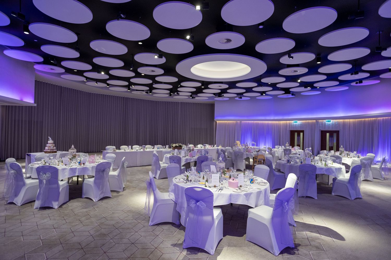 Elegant reception hall with round tables, white linens, and modern ceiling lights; ready for wedding celebration.