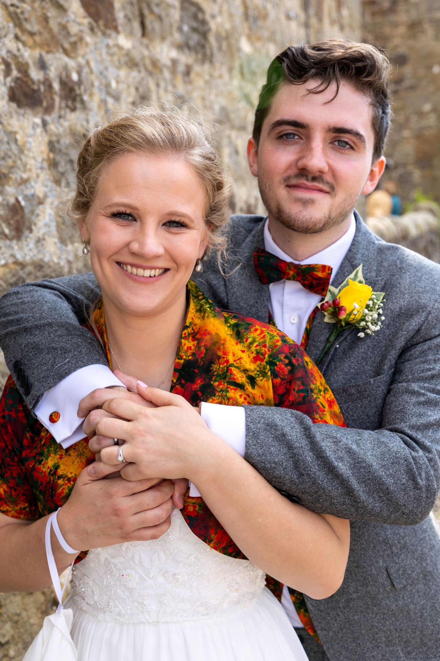 Smiling bride and groom embracing at outdoor wedding, colorful shawl, rustic stone backdrop, joyful couple portrait.