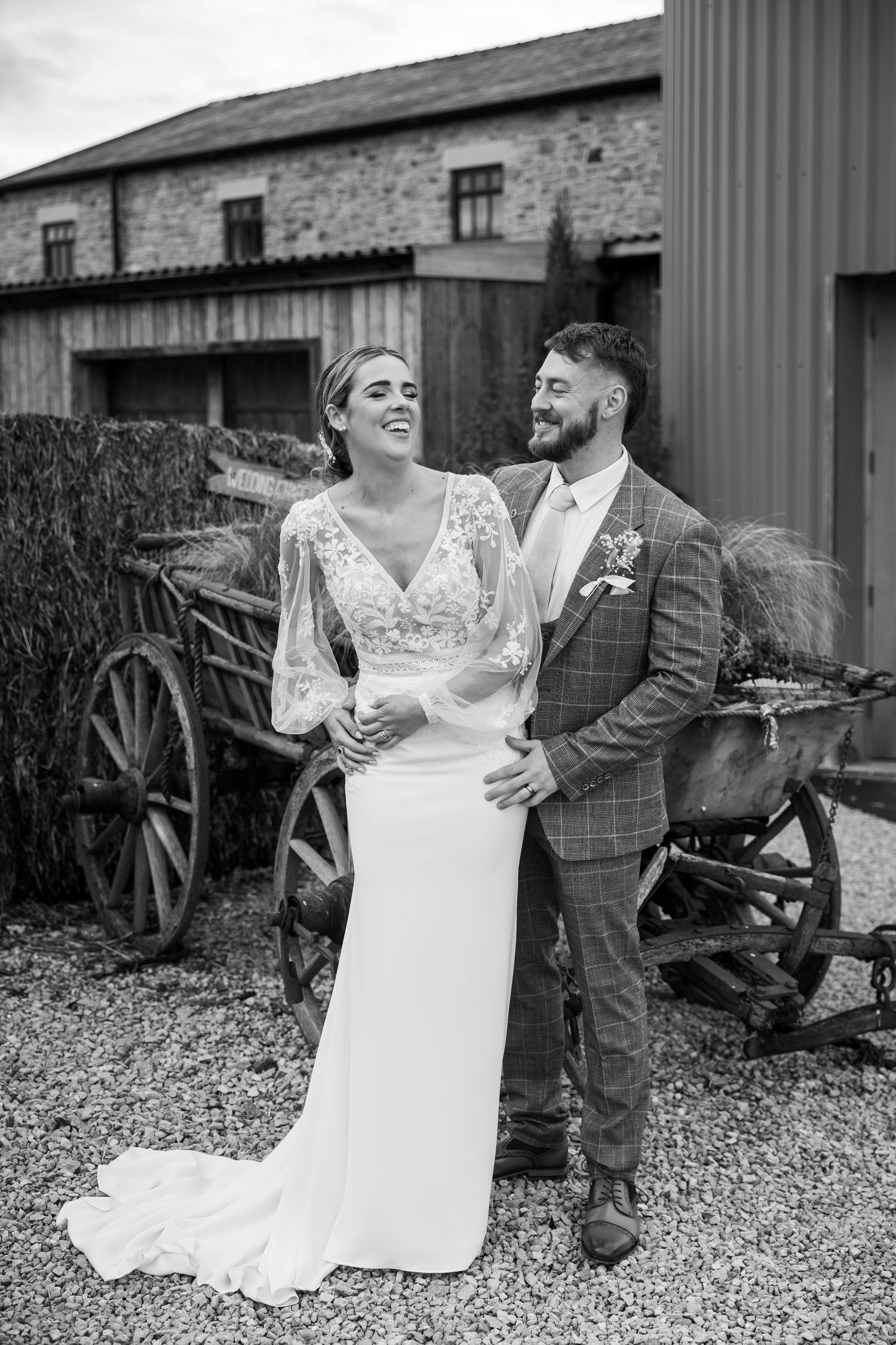 Couple share a joyful moment at Runa Farm; rustic setting with vintage cart and countryside charm in black-and-white.