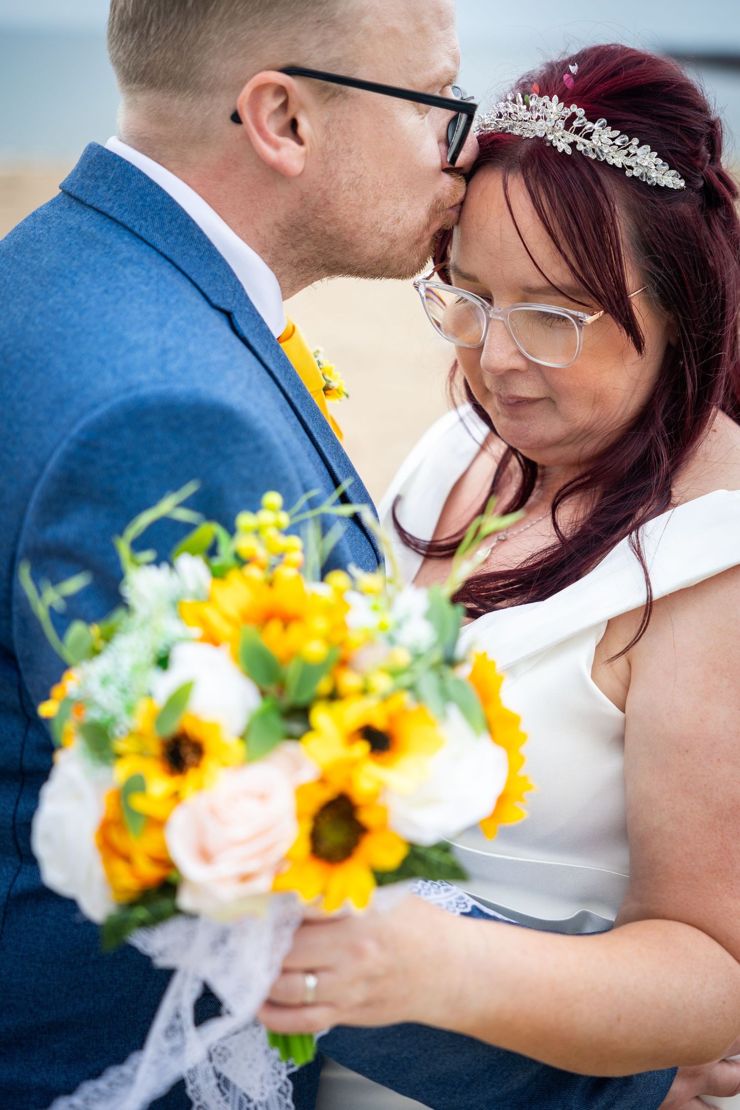 Groom kisses bride’s forehead at Sunderland beach, romantic North East UK wedding moment with vibrant flowers and intimate connection