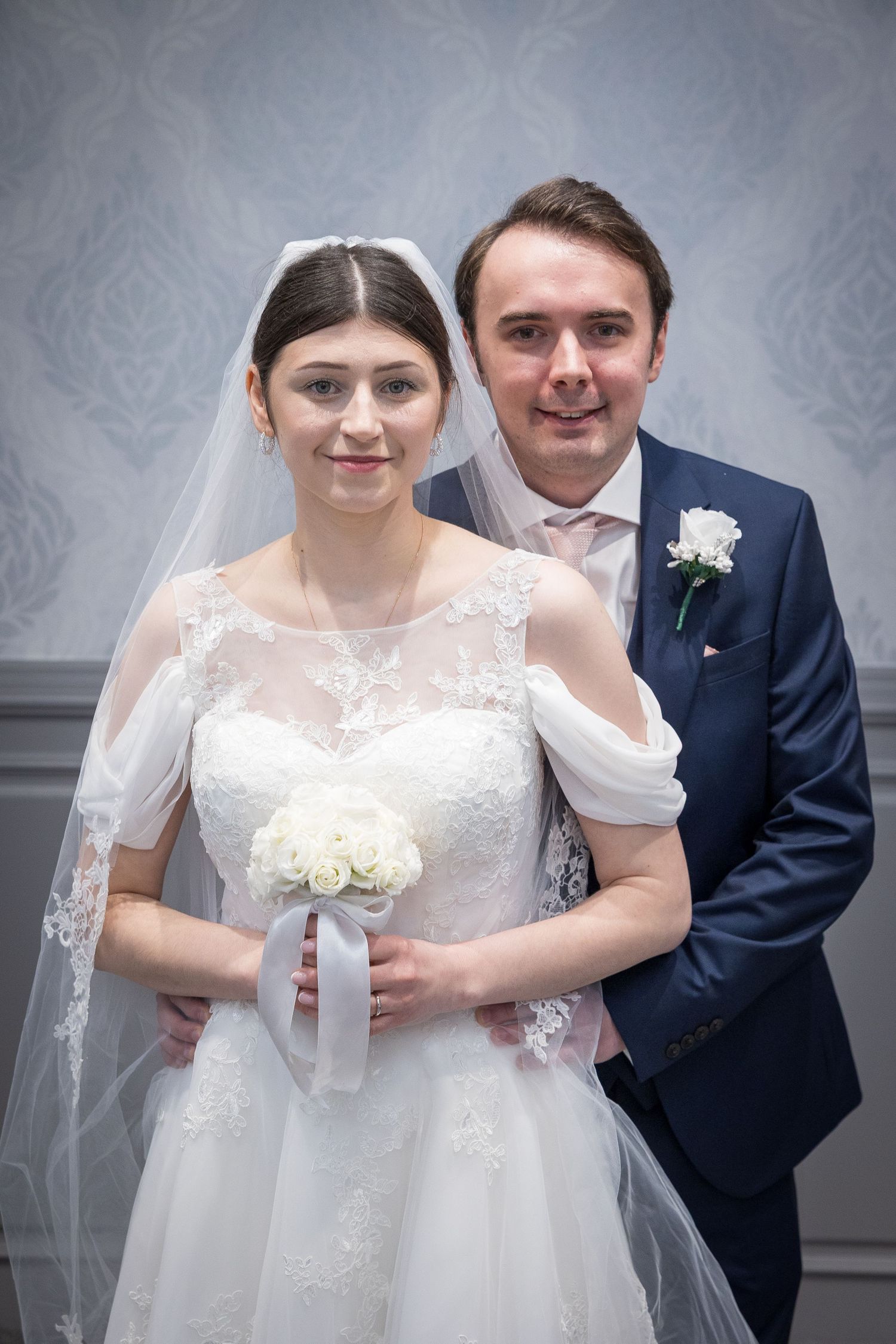 Bride holds bouquet as groom stands behind, both smiling softly; classic posed wedding portrait indoors.