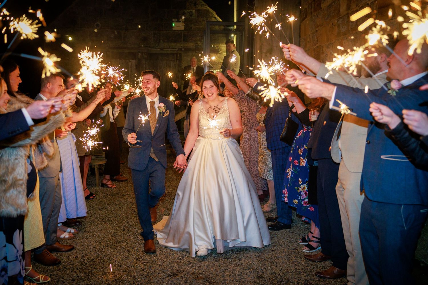 Sparkler exit at Danby Castle; newlyweds walk through cheering guests in a joyful night celebration.