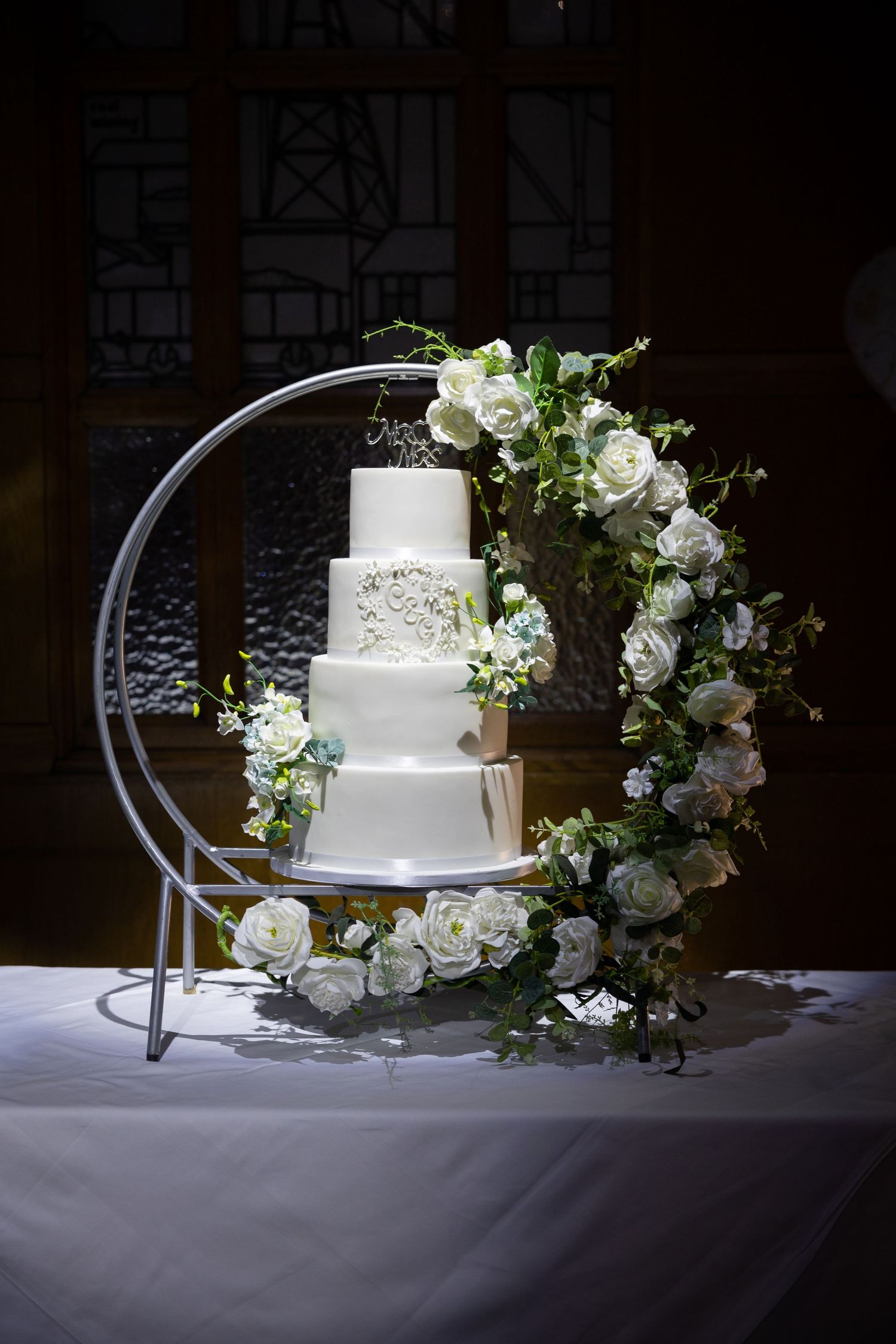 Elegant wedding cake display; three-tier cake framed by floral hoop with soft, romantic lighting.